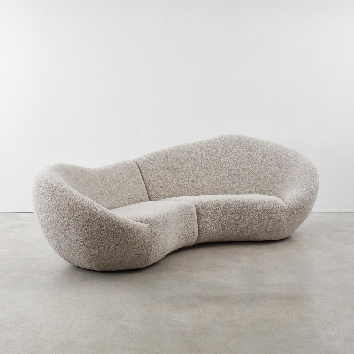 Dramatically sweeping round to envelop the sitter, this late twentieth century curved sofa has an organic form, like that of a bean. The back slopes up asymmetrically for a more supportive back rest on one side. It is formed of two parts and joined