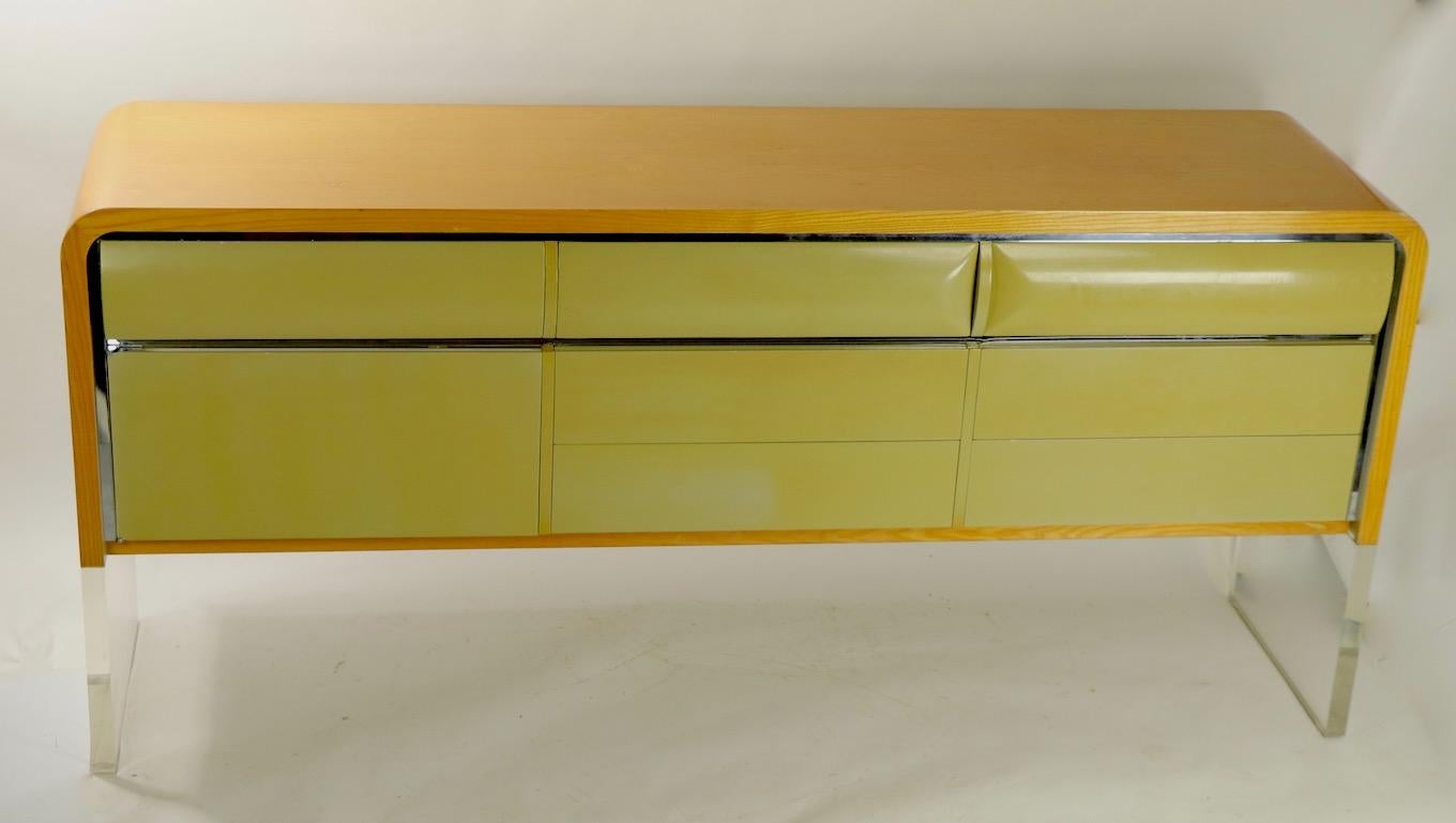 Massive and substantial modernist sideboard having thick (1.5 inch) solid Lucite panel form legs, an oak top with curved waterfall style edges, lacquered drawer fronts and chrome trim. There are 7 drawers which are exposed, and 2 doors which open to