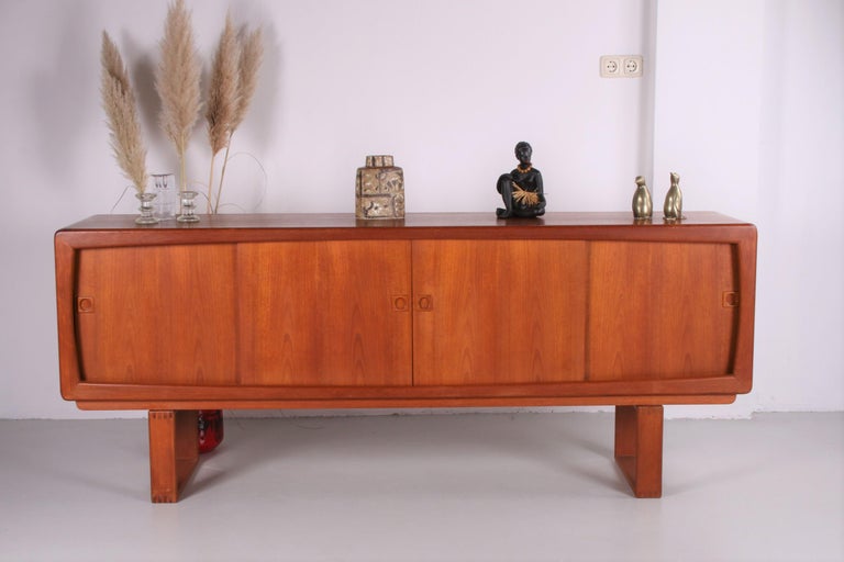This sideboard is made of heavy teak wood. The sideboard itself has a beautiful rounded design with interesting curved legs created by the Danish designer H.W. Klein. There are three compartments and three shelves inside, hidden behind the sliding