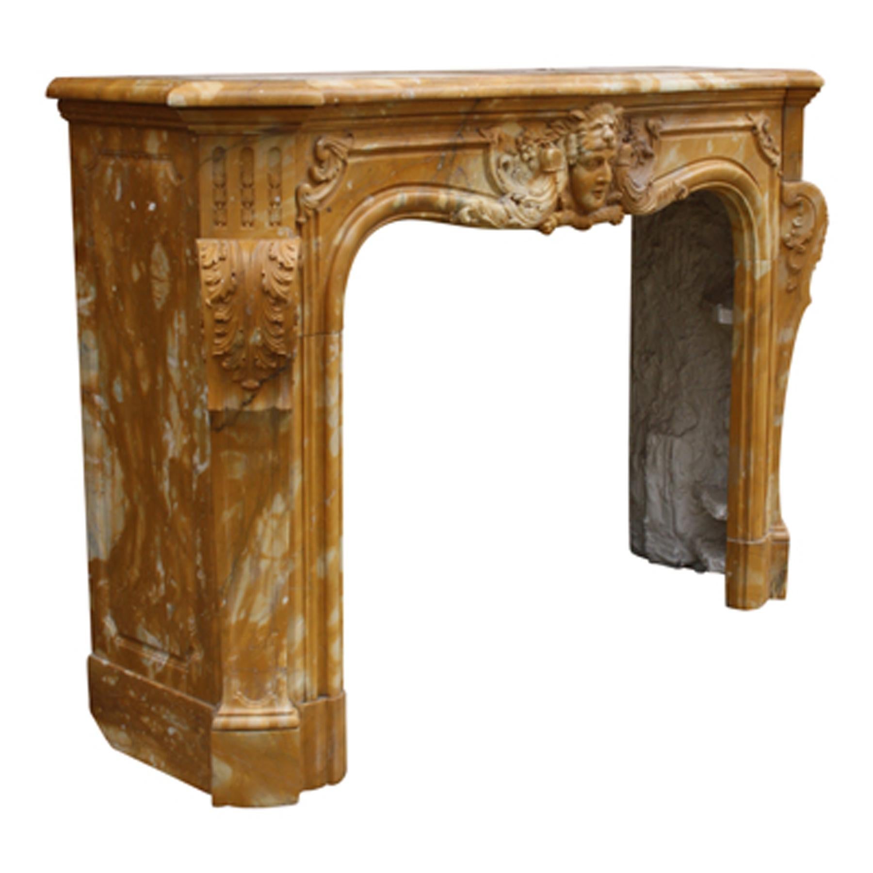 A mid-20th century French Chimneypiece composed of pale Siena marble in the Louis XV manner. The jambs formed of elongated acanthus consoles below triglyph corner blocks above. The serpentine panelled frieze has a central classical mask with a lion