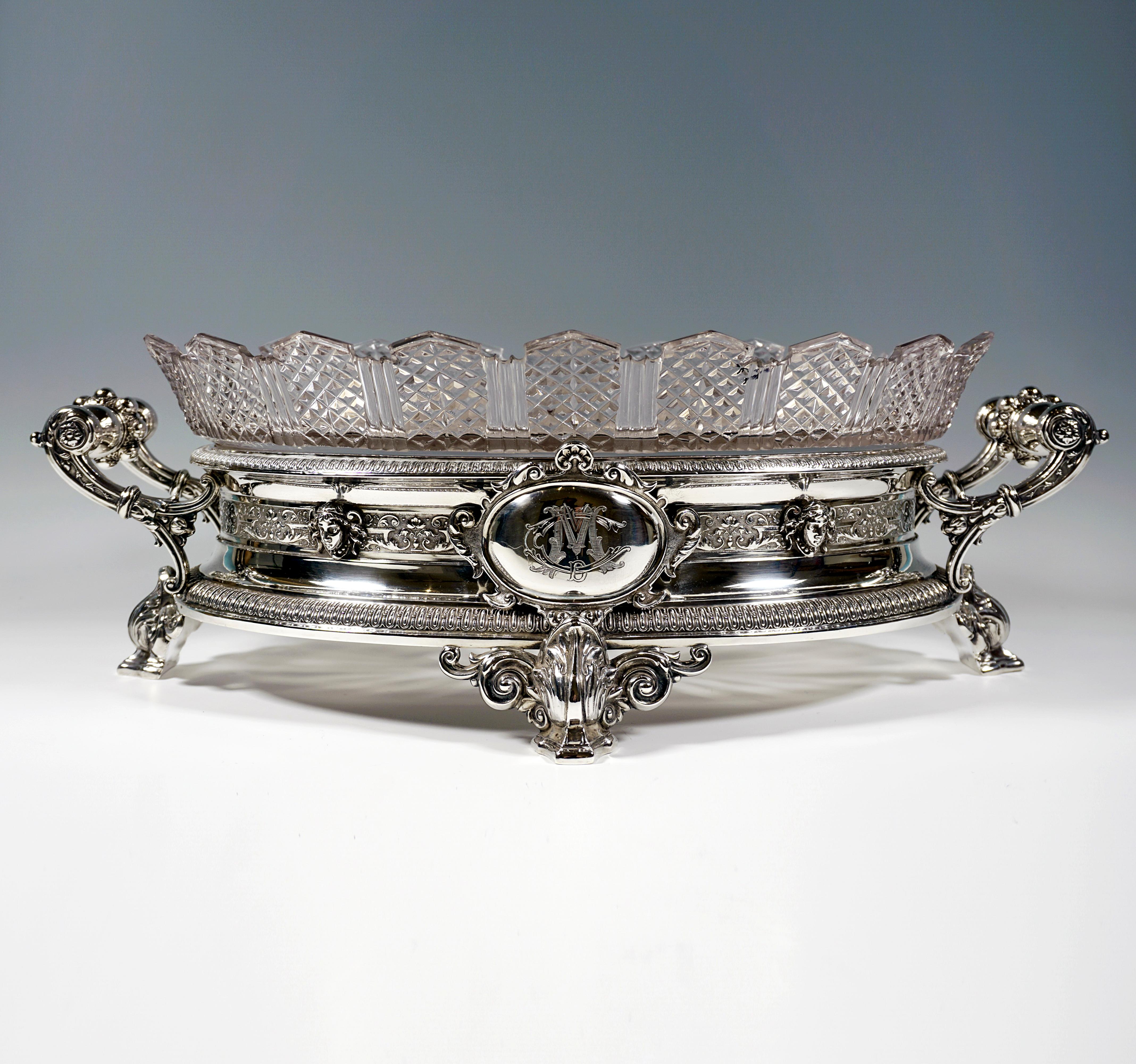 Large festive silver jardinière over oval ground plan, standing on four flared feet with volute decoration and foliate overlays, the body of the vessel bordered on the upper and lower rims by surrounding bands with chased bar decoration, in the