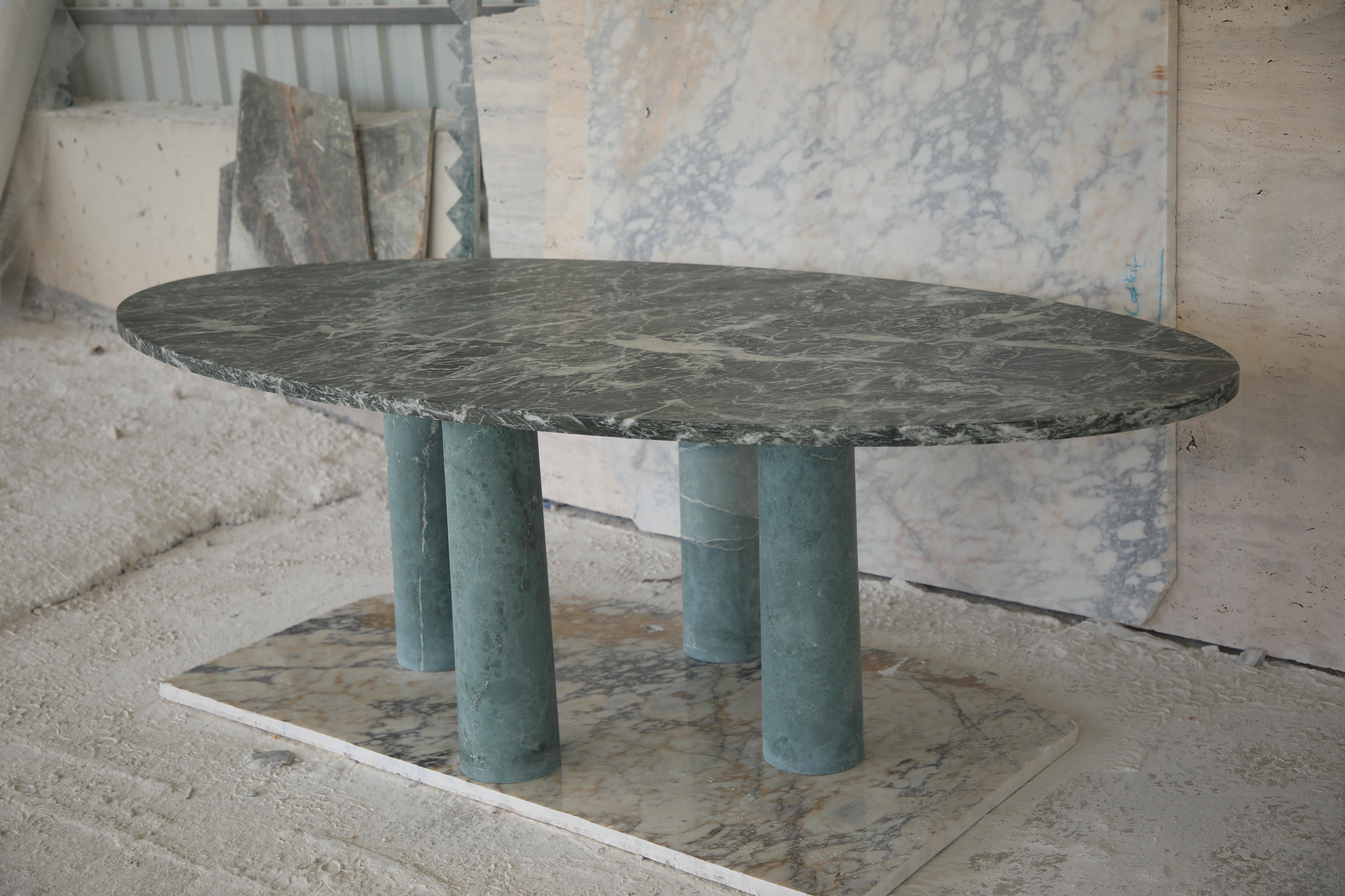 Cette table minimaliste est entièrement réalisée en pierre, avec un plateau ovale en forme de chemin de fer en marbre vert avec d'élégantes veines blanches. Il repose sur quatre colonnes cylindriques qui s'affinent légèrement, conférant à la pièce
