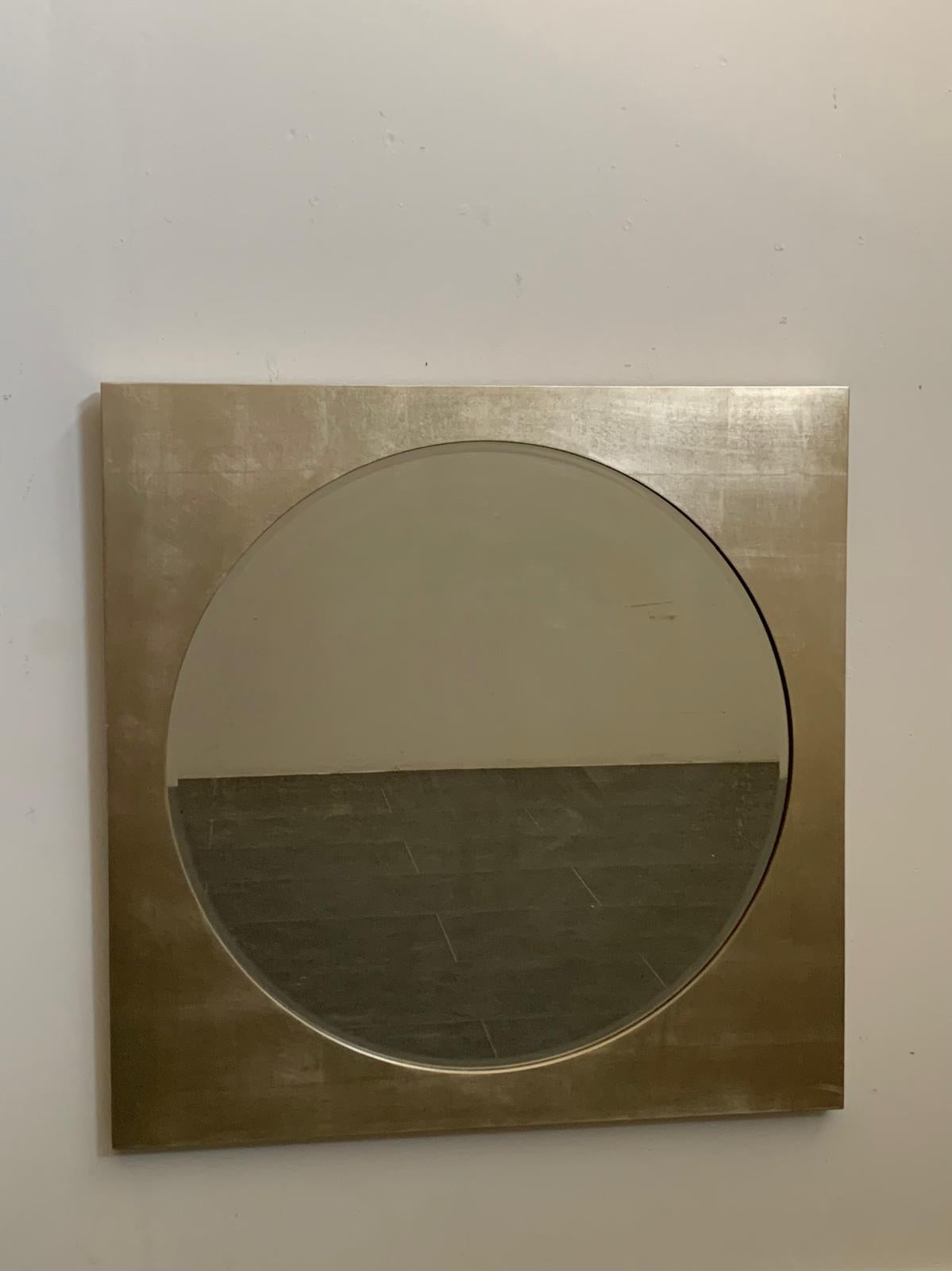 Questo straordinario specchio di grandi dimensioni presenta una cornice quadrata con una sezione circolare centrale che contiene lo specchio stesso. Il design minimalista è esaltato dalla finitura opalescente a foglia d'argento, che enfatizza il suo