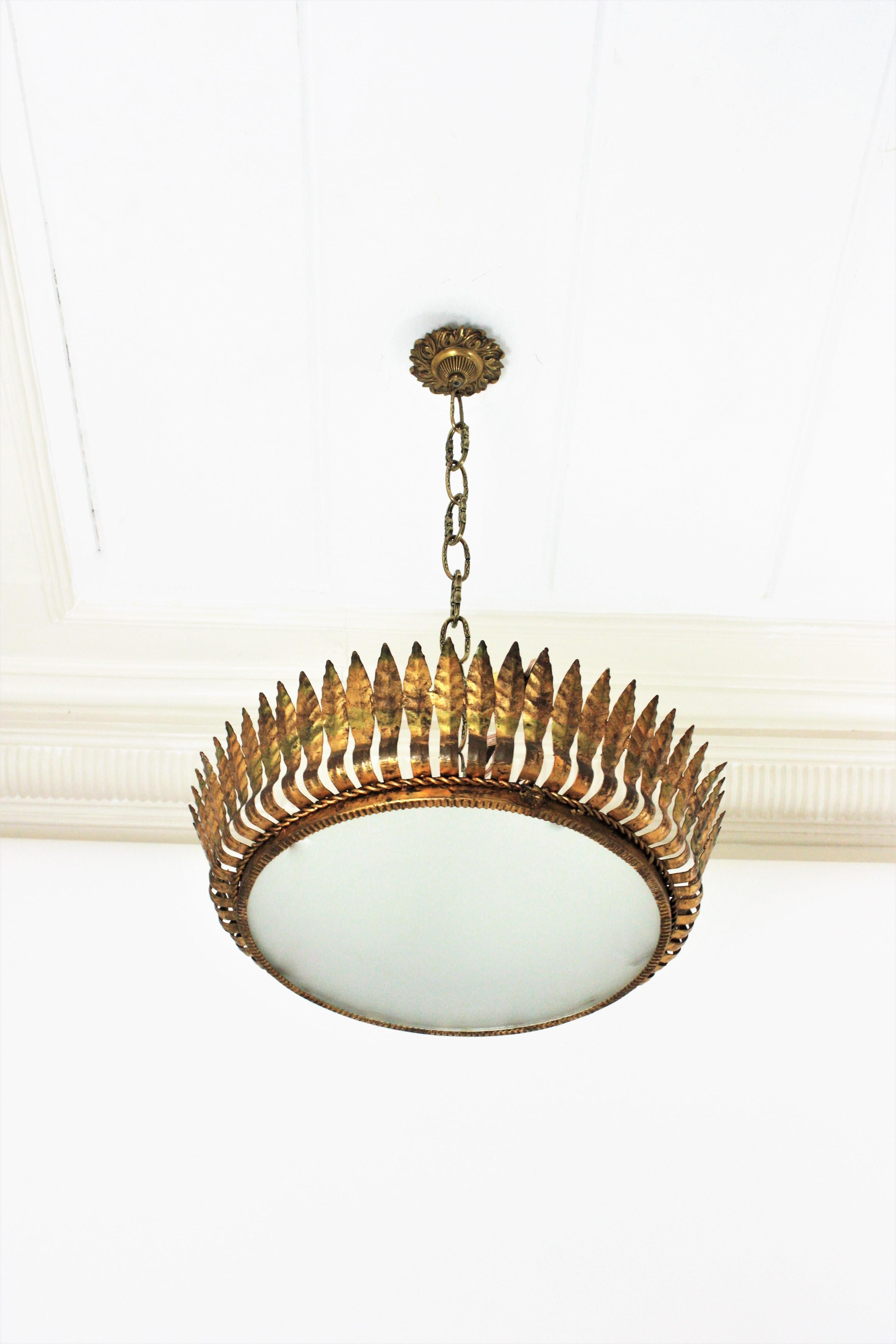 Monture encastrée en forme de couronne, large (20 in ), fer doré. Espagne, années 1950.
Ce luminaire ou cette suspension en forme de soleil présente des motifs de feuilles entourant un diffuseur central en verre dépoli et une corde de fer torsadée