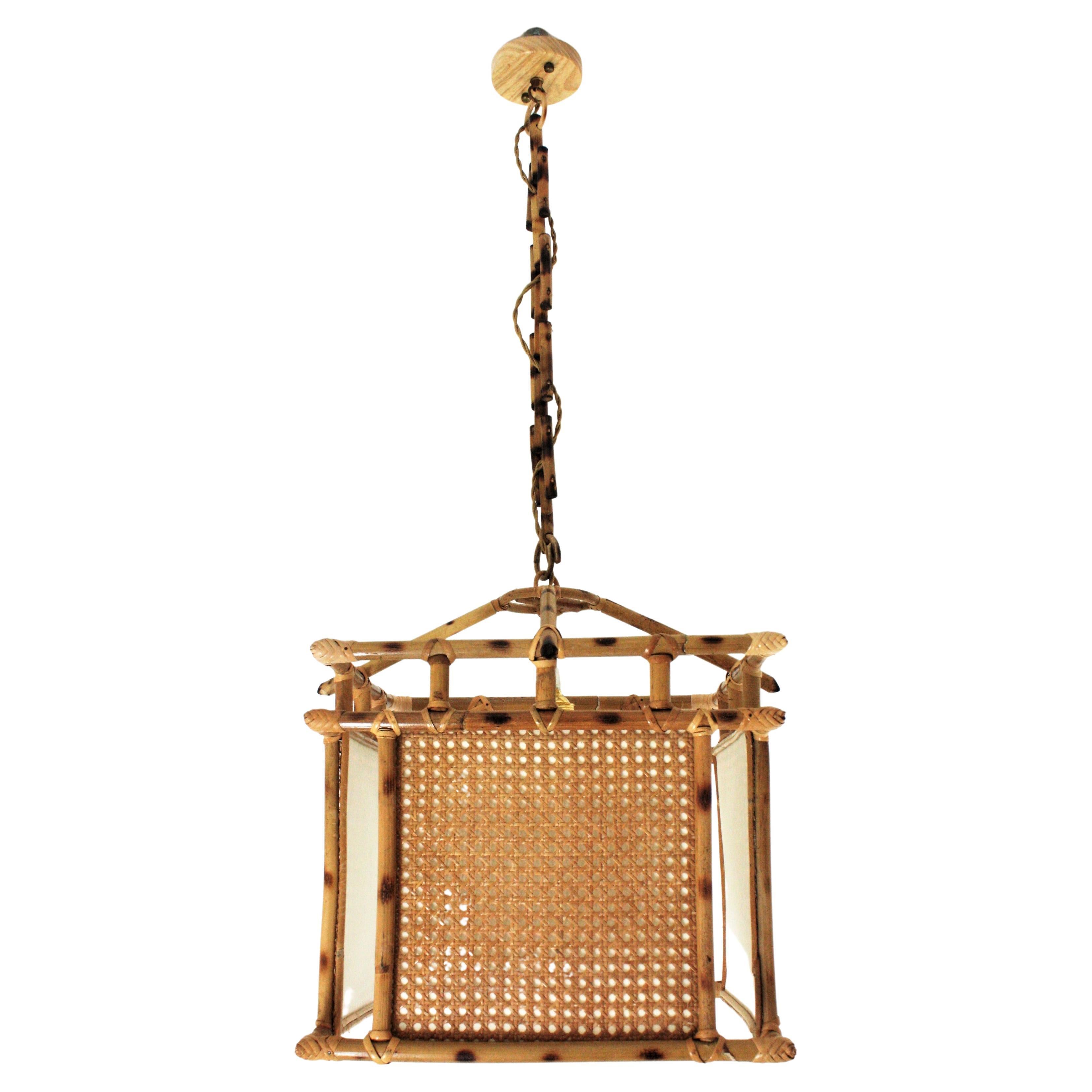 Oriental Inspired rattan squared lantern with wicker wire cannage panels. Spain, 1950s-1960s.
This handcrafted ceiling lamp features a squared lantern made of rattan canes with cannage panels and an inner paper lampshade. The chain is made of wicker