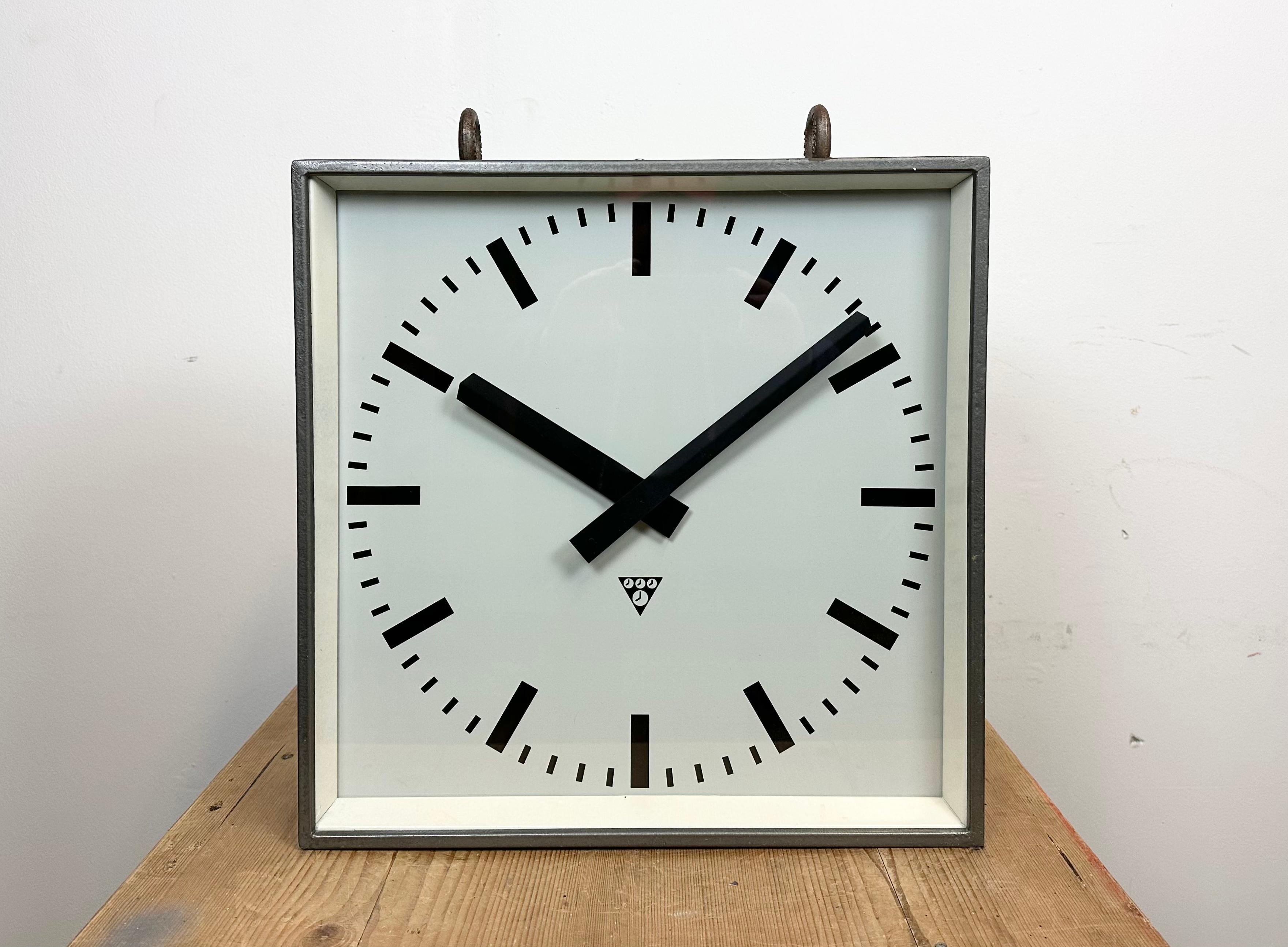 Este reloj cuadrado de doble cara para ferrocarril, escuela o fábrica fue fabricado por Pragotron, en la antigua Checoslovaquia, durante la década de 1970. La pieza tiene un cuerpo de metal gris pintado a martillo, ganchos de hierro, tapas de