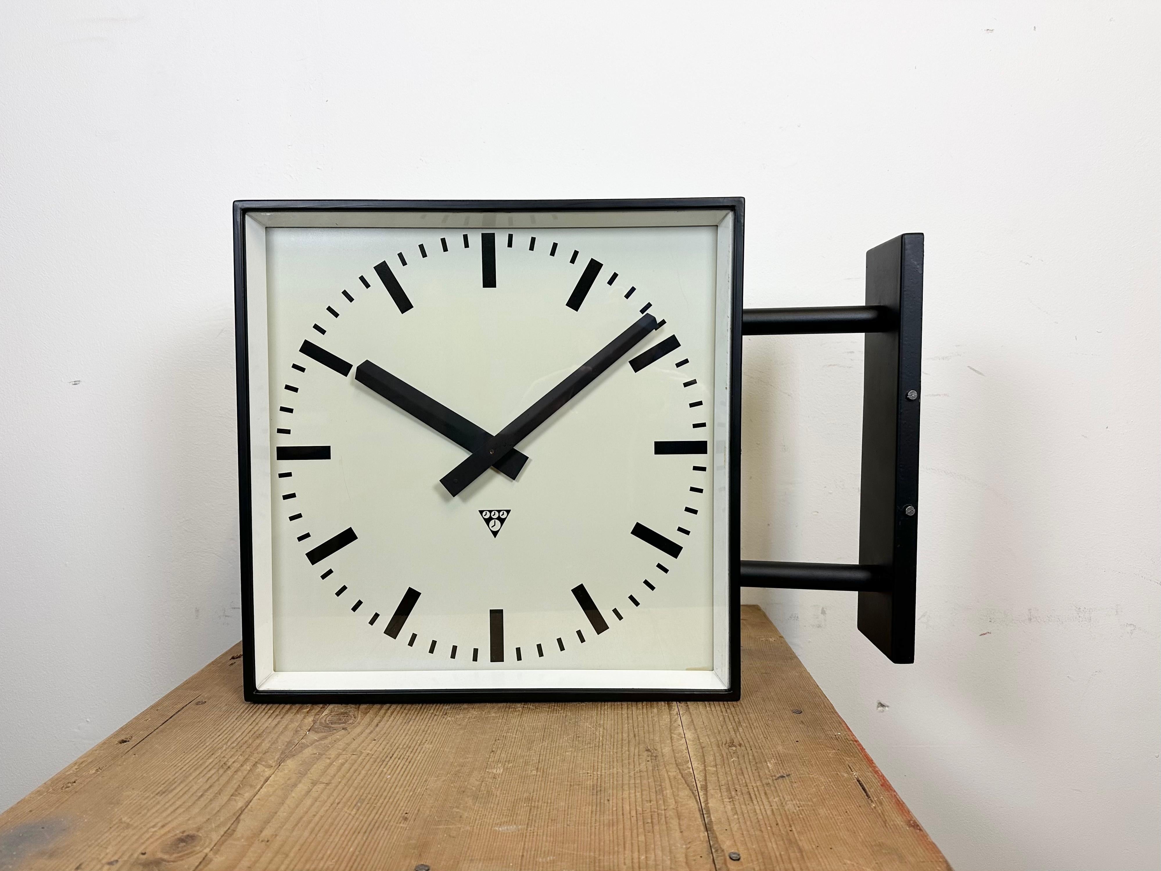 This square double-sided railway, school or factory wall clock was produced by Pragotron, in former Czechoslovakia, during the 1970s. The piece features a black metal body, a clear glass cover, and an aluminum dial and hands. The clock has been