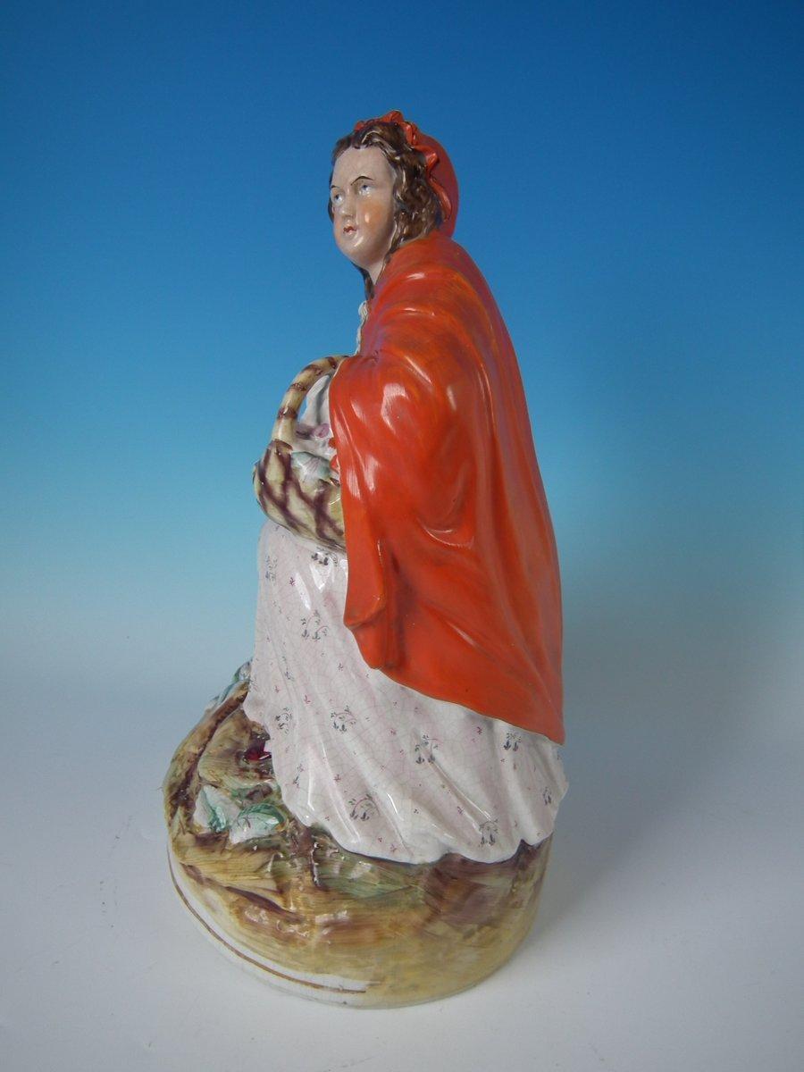 Staffordshire pottery figure with a theatrical and literary theme which features red riding hood, with a wolf beside her, seated on a circular base. Dull gilt base line and embellishment. Decorated 'in the round' - decoration to front and reverse.