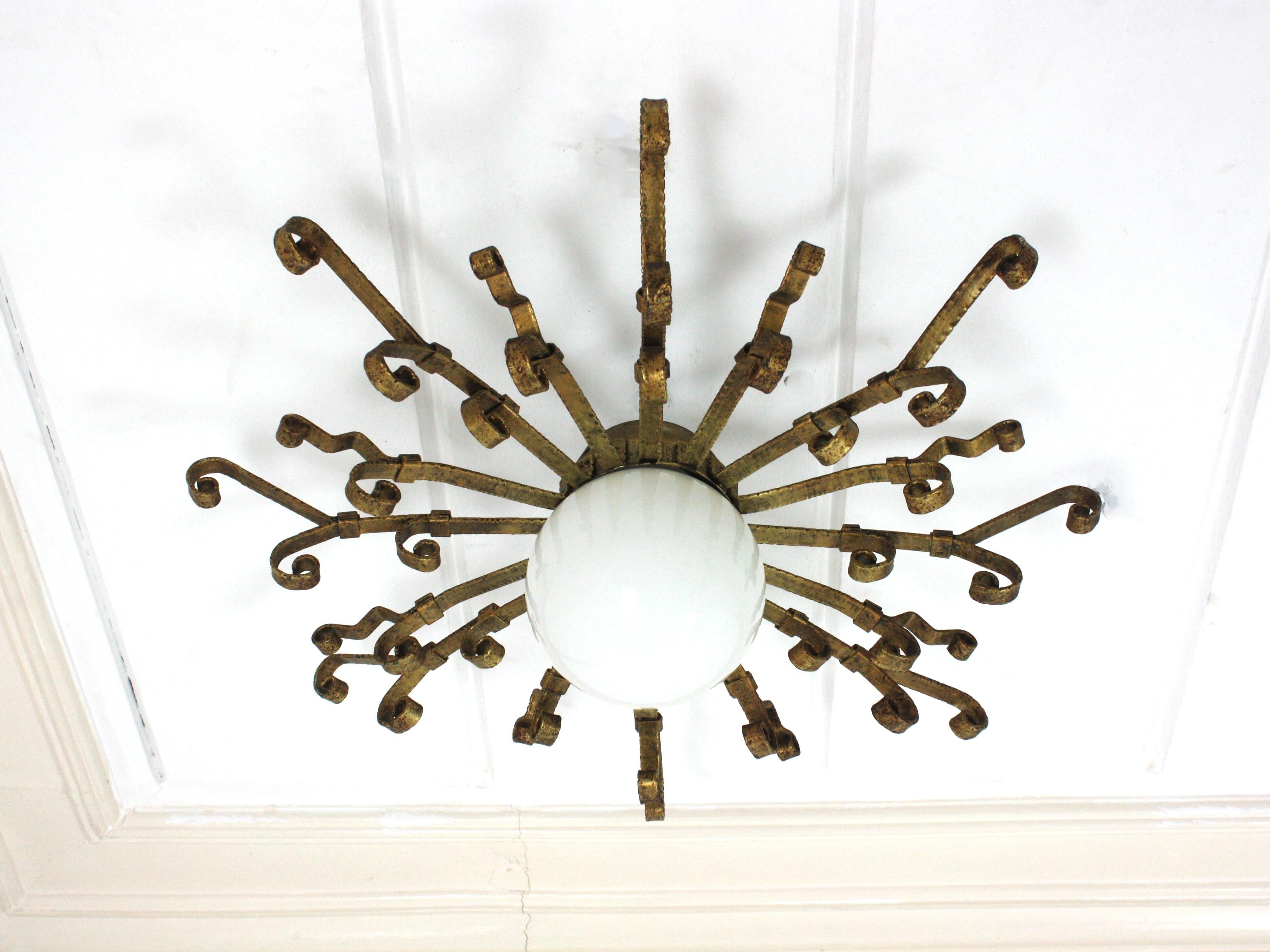 Eye-catching gilt wrought iron sunburst ceiling flush mount with milk glass lampshade and scroll work accents. Spain, 1950s
Rare find. 
This light fixture features a sunburst structure with scrollwork rays made in hand forged iron surrounding a