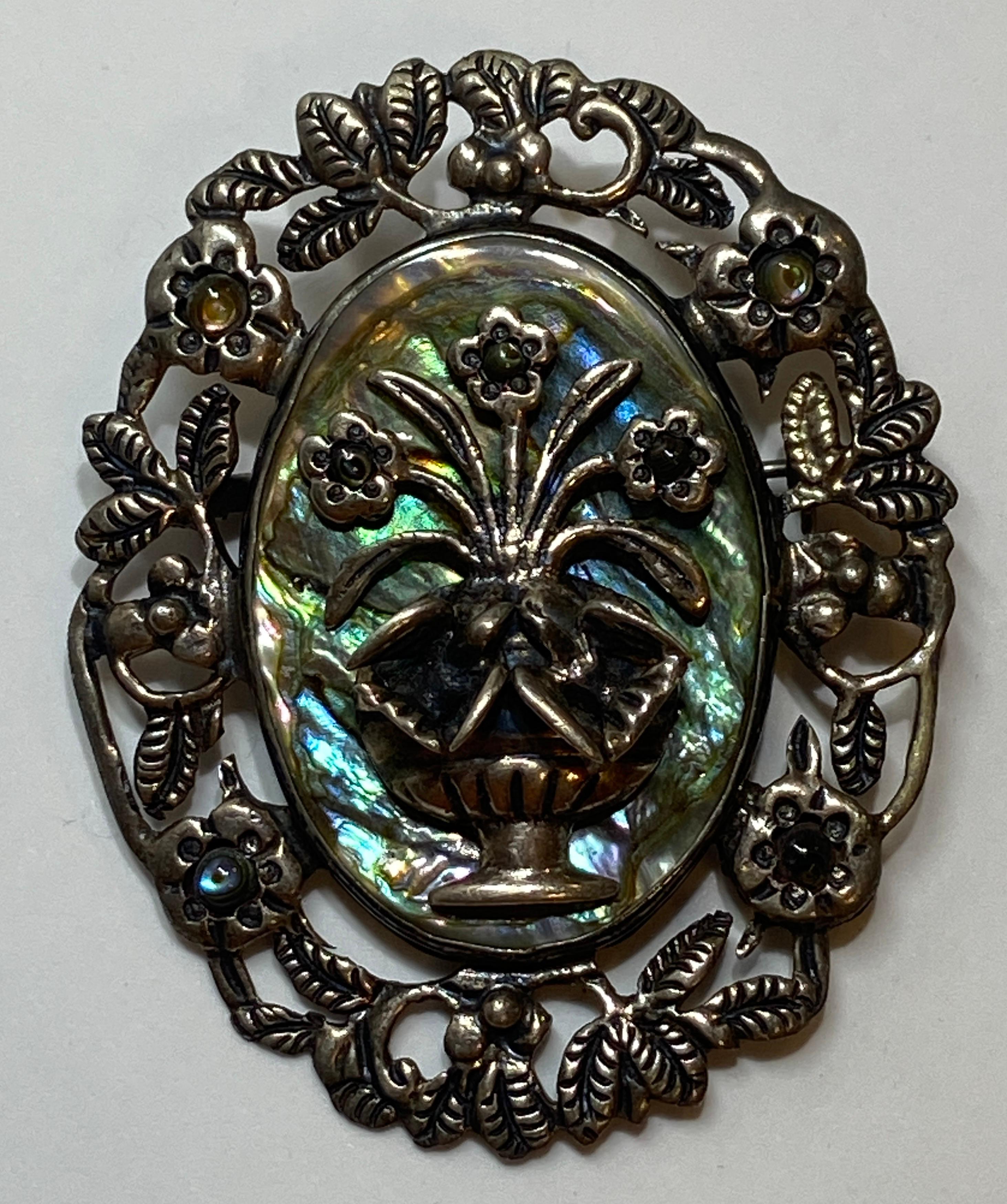 Wonderfully detailed large brooch accented with a abalone center back-drop for the centerpiece featuring a 3-dimensional 'Vase with Fresh-Cut Florals'. Brooch is bordered with floral leaves and florals. This sterling silver brooch has the 'maker's