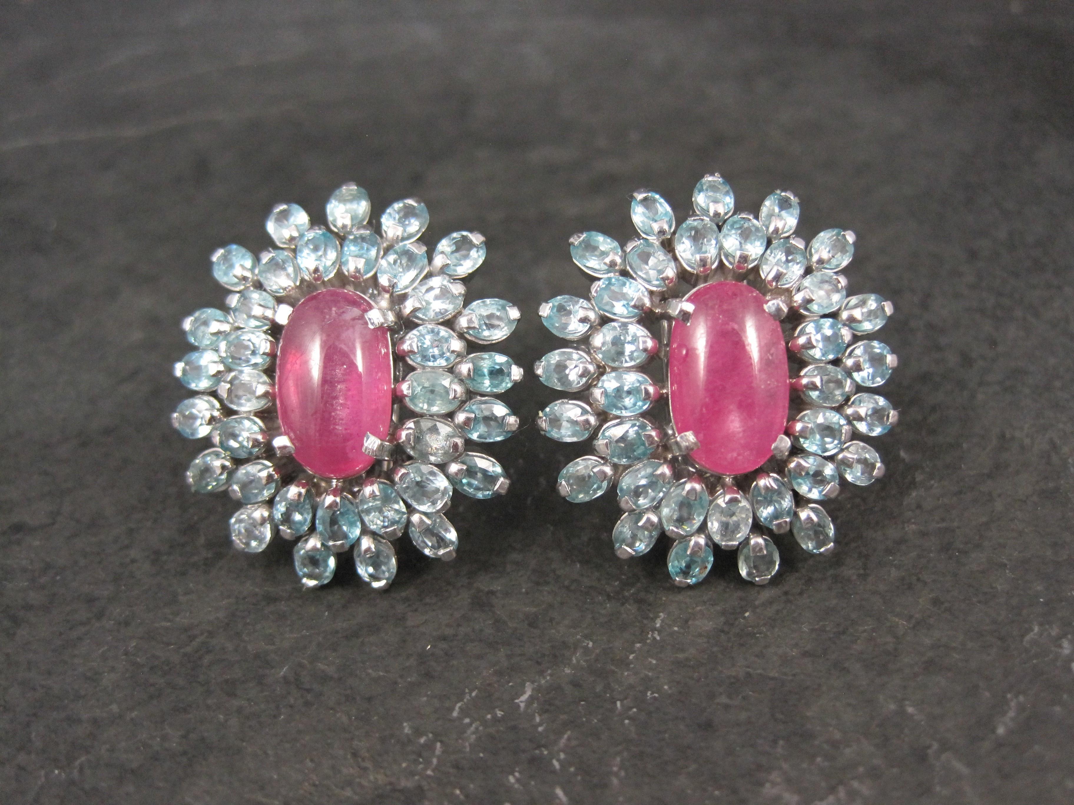 These gorgeous estate earrings are sterling silver with French backs.
They feature 14x9mm cabochon cut pink sapphires accented by a halo of 4x3mm blue topaz gemstones.

Measurements: 1 1/8 by 1 1/4 inches
Weight: 24.4 grams

Marks: 925

Condition: