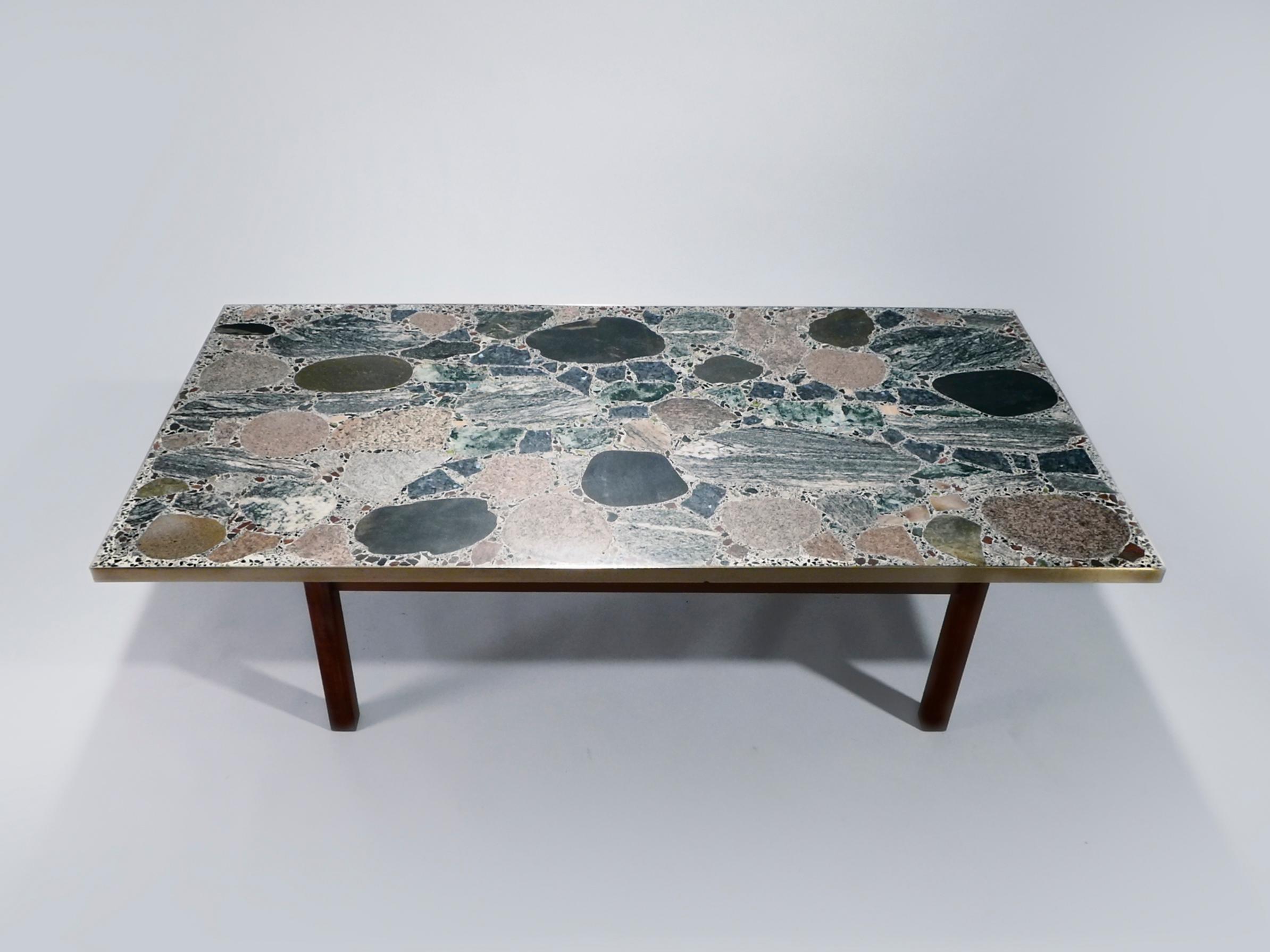 The thick surface of this coffee table is intriguing, created from heavy, multicolored stones. The result is a sophisticated, naturalist look that is complemented by strong, unadorned teak feet and brass frame. Chic, Scandinavian style is