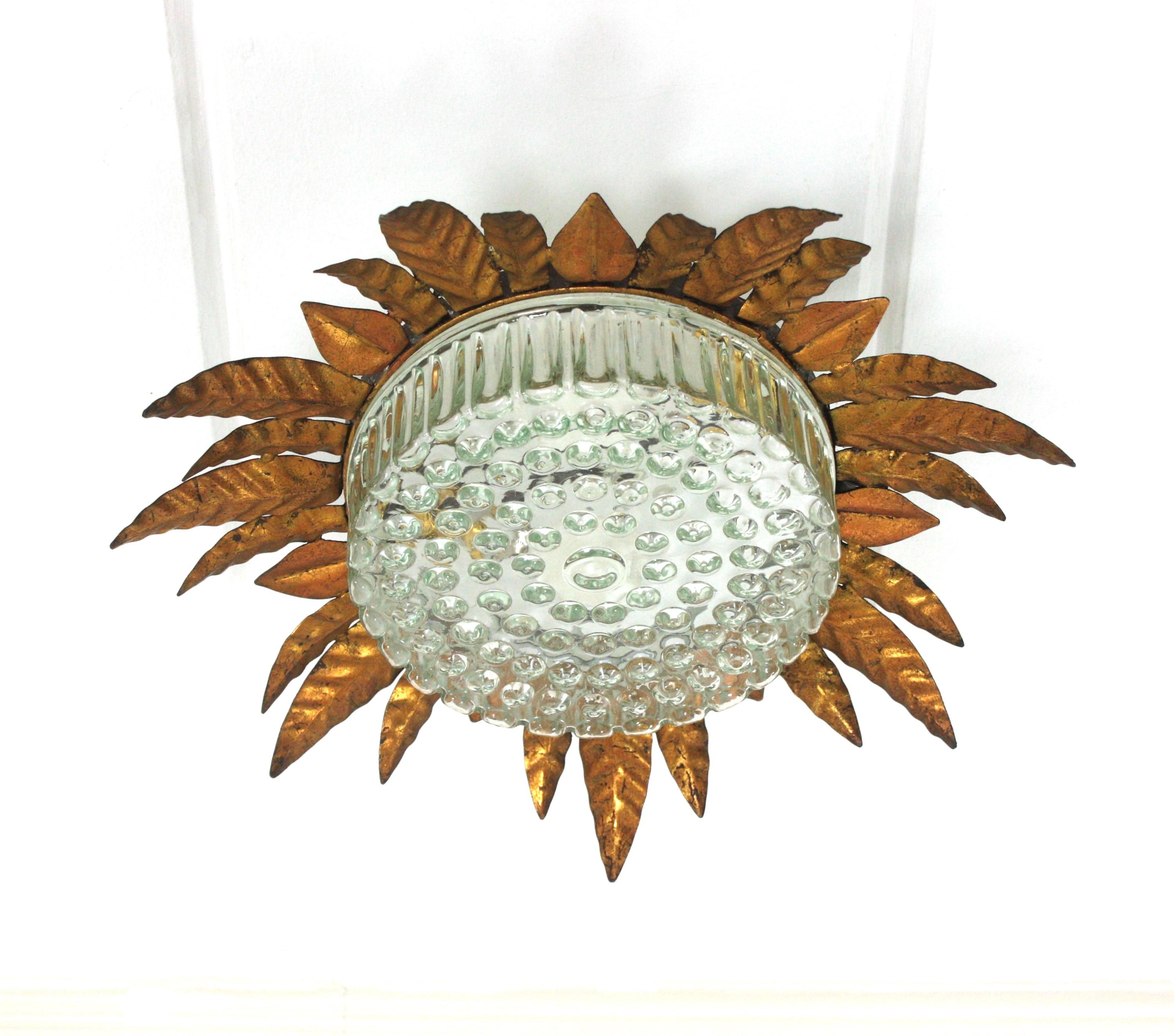 Large Flower shaped foliage sunburst flush mount, Gilt Metal, Pressed Glass, Spain, 1950s.
This ceiling lamp features a gilt iron leafed frame with alternating leaves in three sizes. It surrounds a central pressed glass shade with bubble pattern