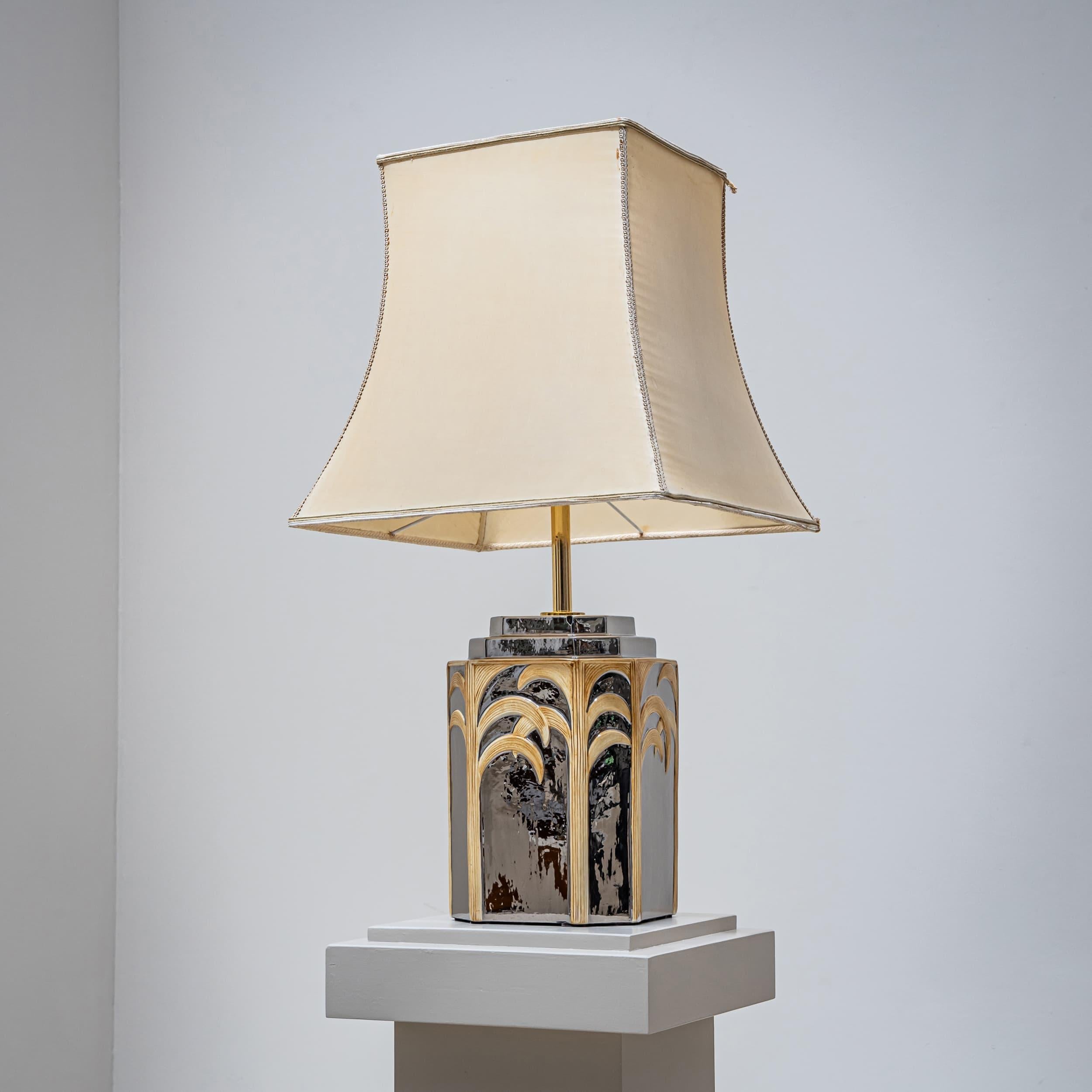 Large table lamp with a beige fabric shade and a ceramic base with silver overlay, decorated with stylized palm motifs in beige.