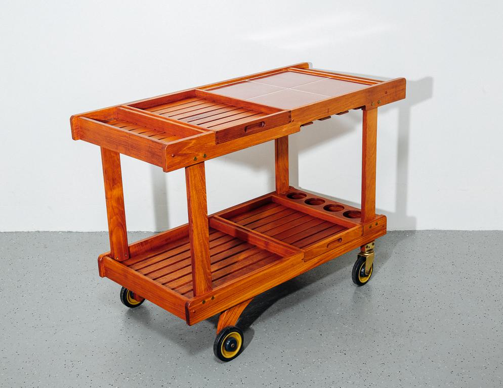Teak slat bar cart with red tile top and removable serving trays. Stem glass storage on underside of top. On casters.