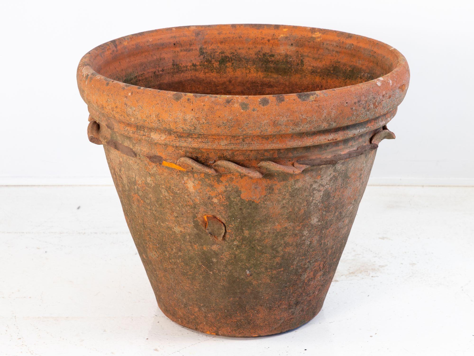 Un grande vaso antico in terracotta con un ampio bordo svasato e un dettaglio decorativo a corda che circonda la parte superiore. La superficie ha sviluppato una ricca patina grazie al muschio naturale e agli agenti atmosferici, che ne esaltano il