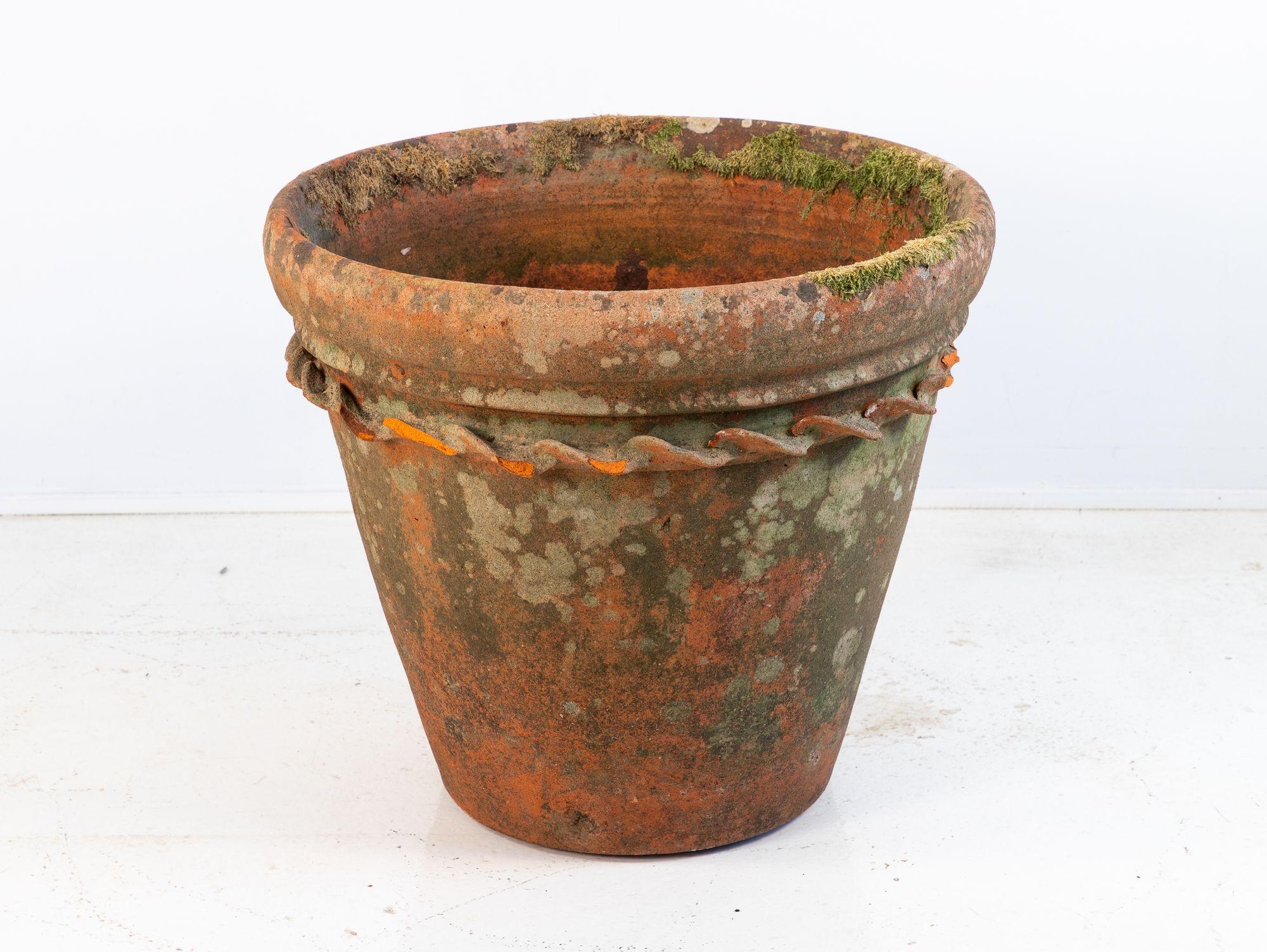 A large antique terracotta pot featuring a wide flared rim and a decorative rope twist detail encircling the upper body. The surface has developed a rich patina with natural moss and weathering, enhancing its rustic character and authenticity.