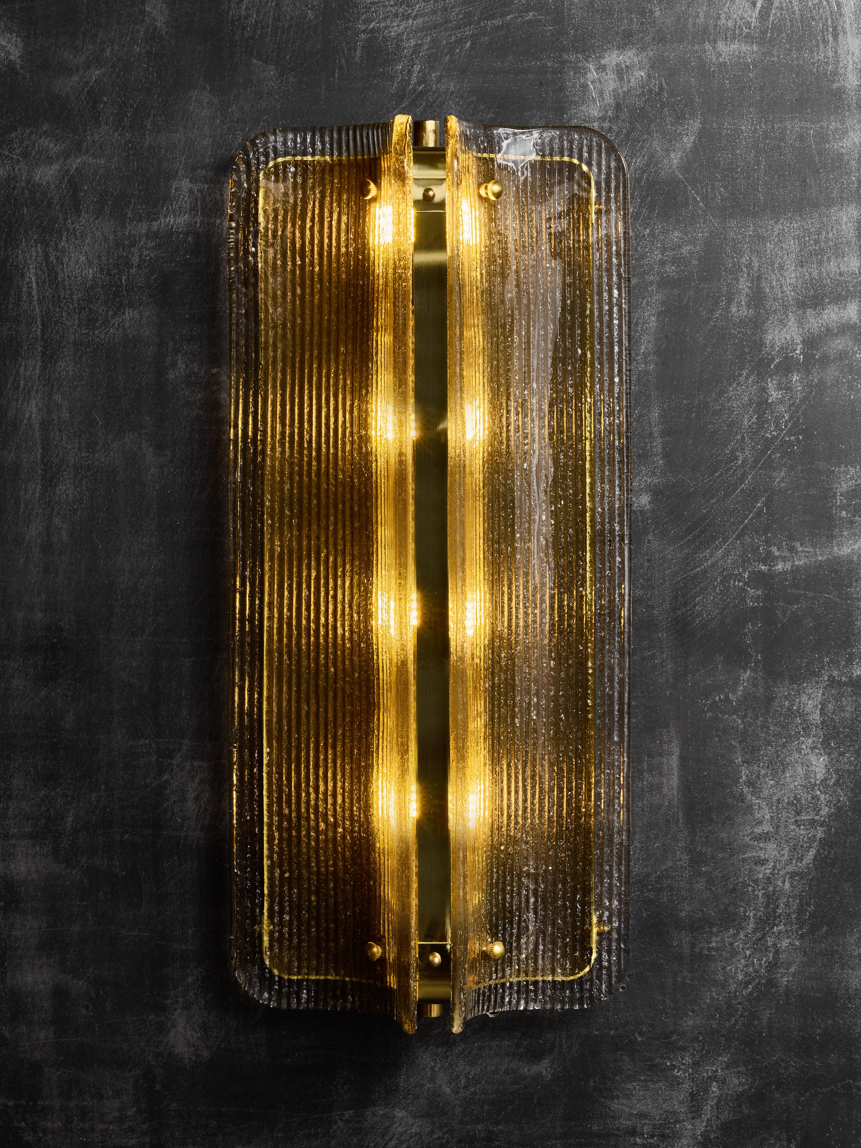 Un sofisticado aplique de pared moderno, con un conjunto escultural de paneles de cristal texturizado montados alrededor de una estructura central de latón. La lámpara se compone de dos elementos de cristal curvados verticalmente, cada uno con una