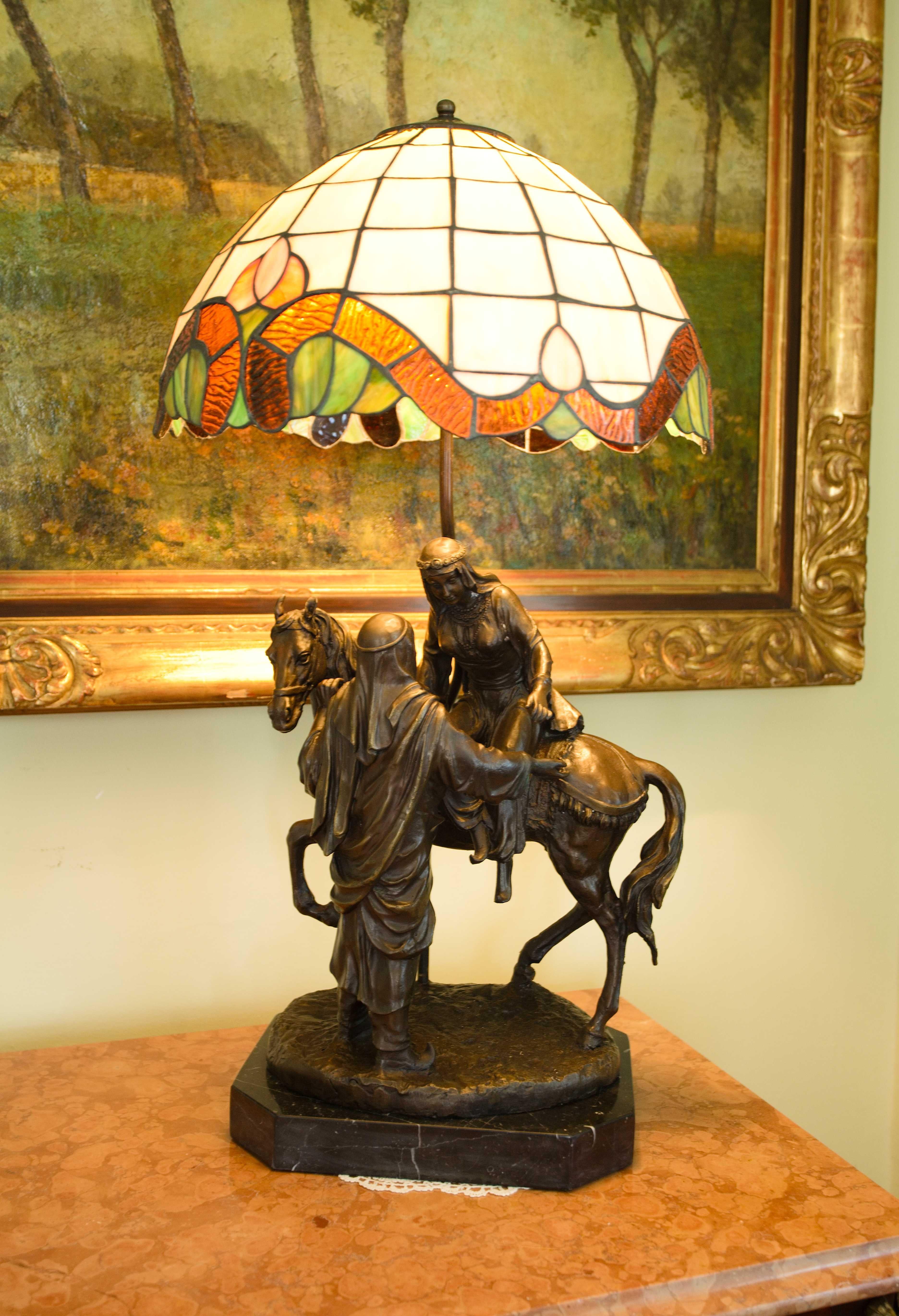 Large Tiffany table lamp. At 75 cm

A beautiful Tiffany style lamp with a colorful glass shade. The body of the lamp is made up of a sculpture depicting a man helping a woman to dismount from a horse. The lamp is signed and has a mark - see photo.
