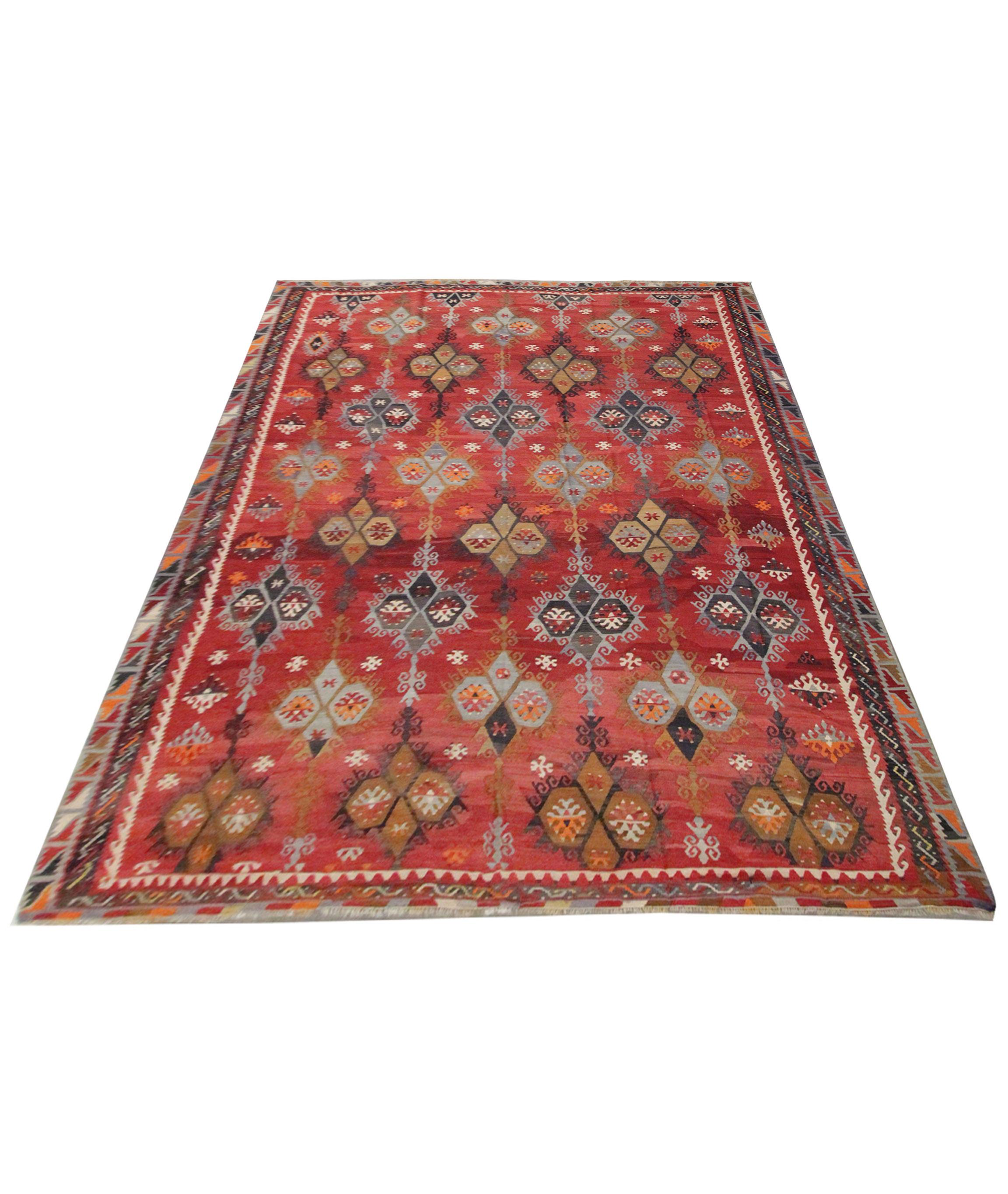 This bold rust-red rug is a traditional tribal Kilim woven in the early 21st century. The design has been woven with symmetrical tribal motifs in mustard, grey and blue accents. A bold repeating pattern layered border then encloses this. The colour