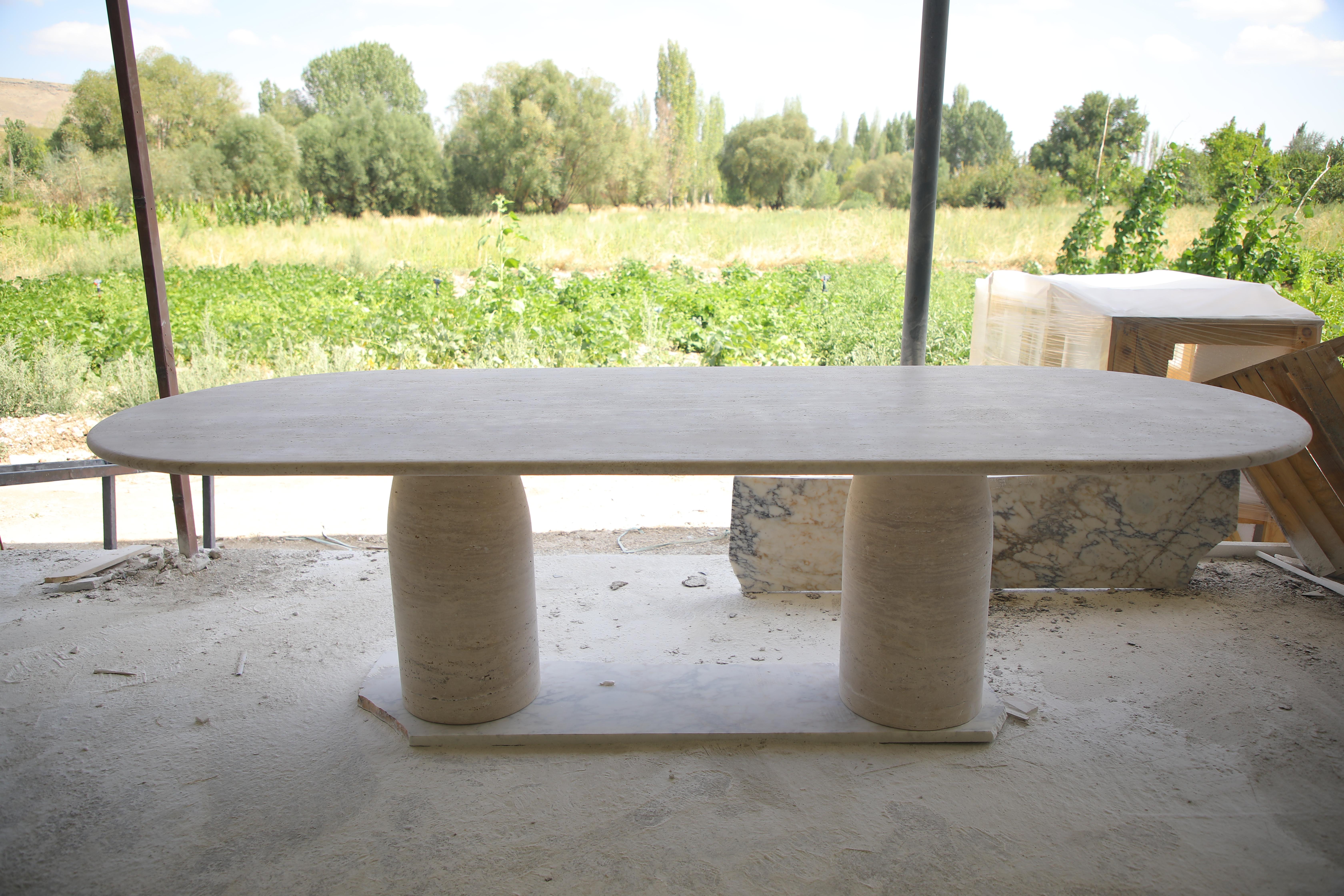 This minimalist table is crafted entirely from a single material, showcasing a beautifully curved-edge top in natural travertine. It rests on distinctive bullet-shaped legs, strategically positioned to comfortably seat 8 to 10 people. The honed
