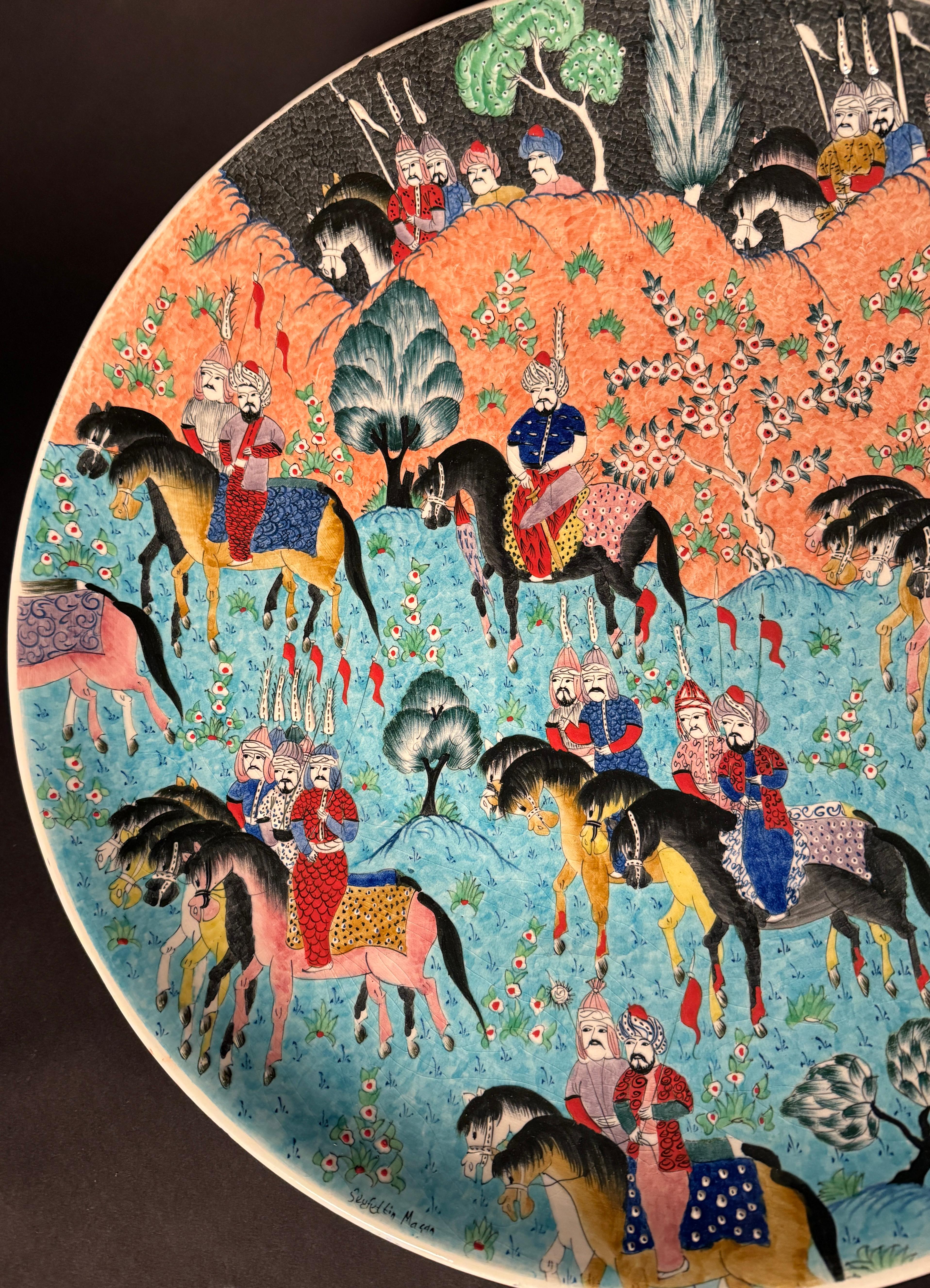 Gran cuenco/cargador de cerámica turca pintado a mano.  Procesión o cacería real en la que participan hombres a caballo vestidos con trajes tradicionales que portan banderas de procesión/desfile. Es un retrato muy detallado, con colores vibrantes y
