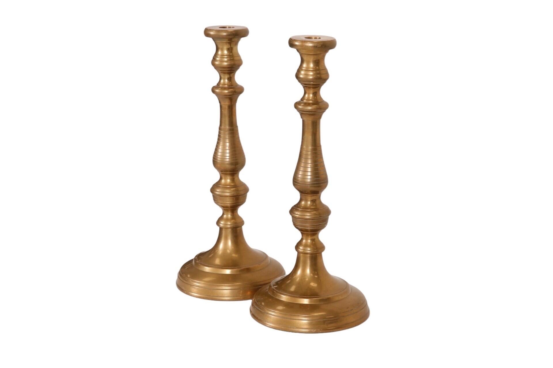 A large pair of turned brass candlesticks. Recessed sockets sit atop turned capitals and columns on round bases. Dimensions per candlestick.
 