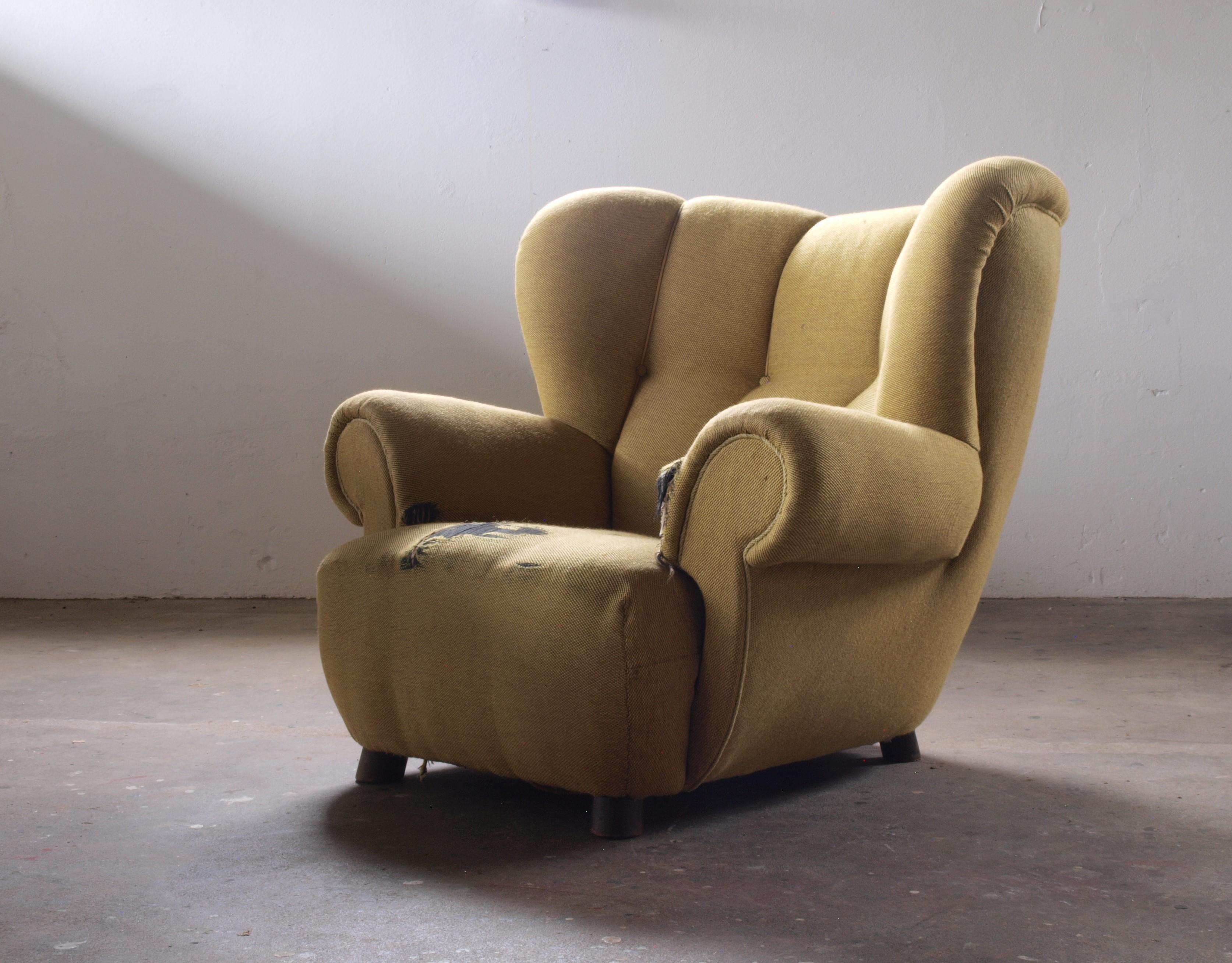 Large upholstered armchair from around the 1930s. Comfortable and embracing. Comparable to Flemming Lassen's style. Potentially reupholstered in lambswool. Requires ample space due to its size and presence. Comes with a matching footstool for