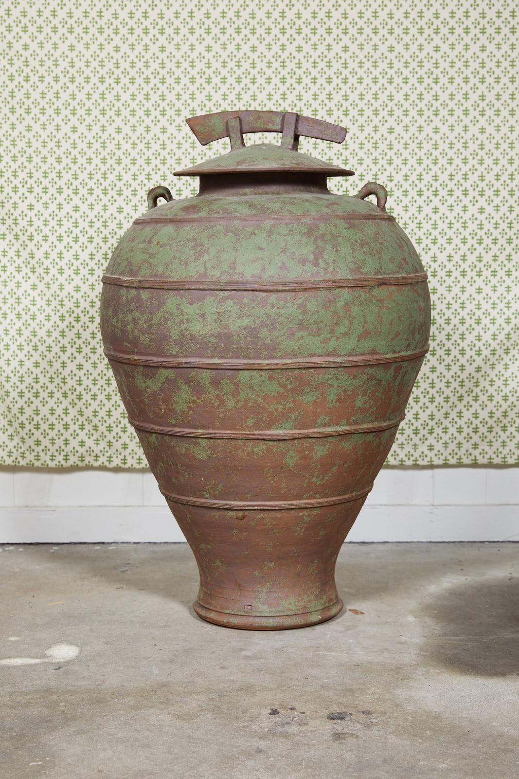 Large urn shaped clay jar with ornamental lid having a bronze and green patina and stamped by the maker. The jar has a pair of handles near the mouth of the vessel, attractive horizontal ribbing, and a decorative drain hole at it’s tapered base.