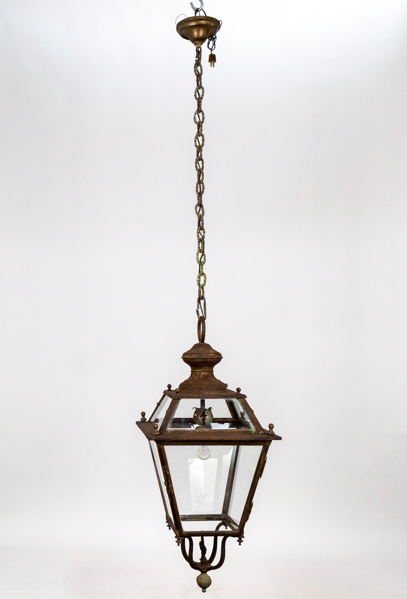 A large, tapered, 4-sided, antique hanging lantern with a beautiful patina texture in bronze and rust. It has a twisted-link chain with some verdigris in the variegated patina and leaf details on the socket cover and a large fleur de lis on top of
