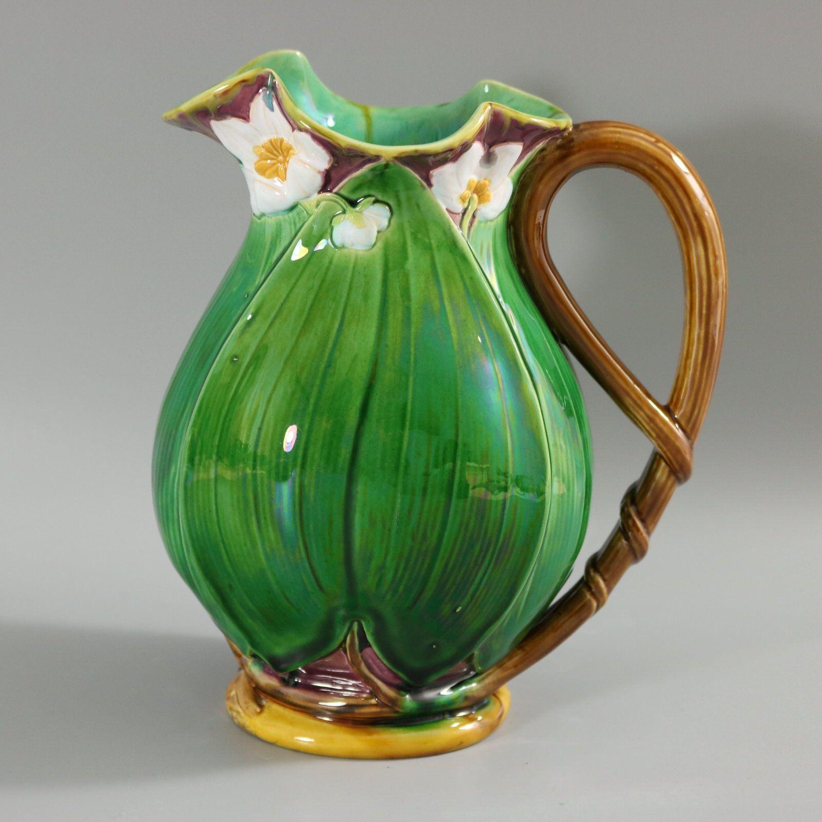 Minton Majolica jug/pitcher which features flowering lilies and lily pads. Colouration: green, white, dark pink, are predominant. The piece bears maker's marks for the Minton pottery. Bears a pattern number, '1228 12'. Marks include a factory