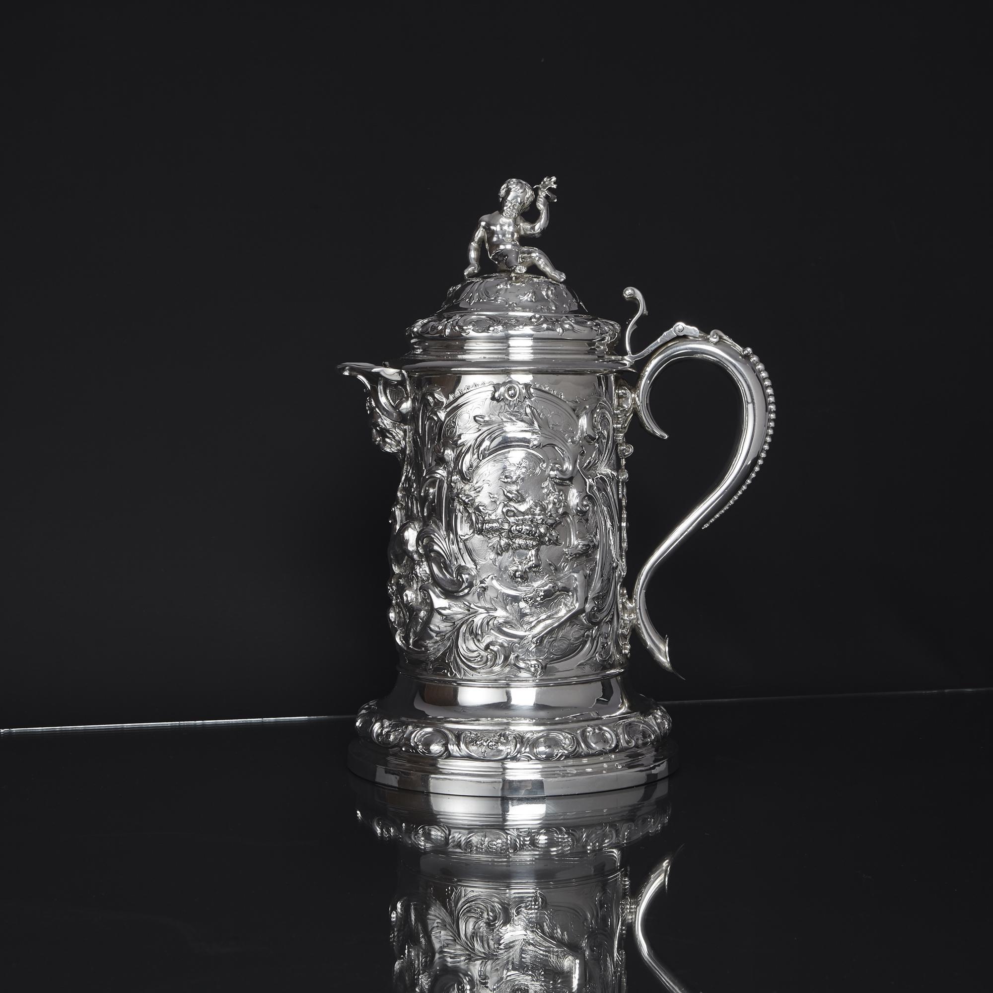 An impressive silver beer jug with high-relief, chased scenes of a stag hunt among scrolling foliage and floral designs. This substantial piece, made by a fine silversmith, has a cast mask spout and hinged lid featuring a charming cherub finial and