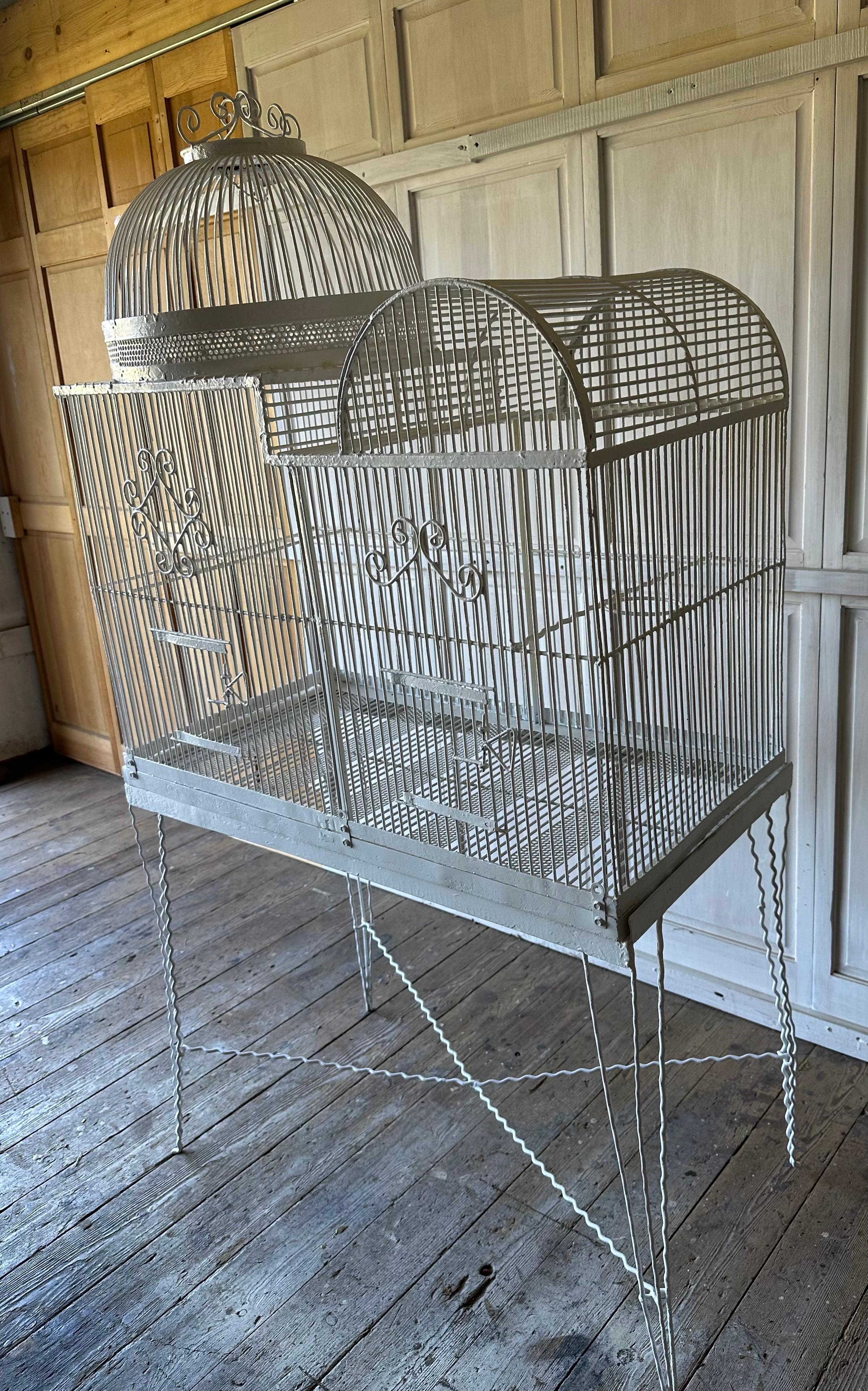 Una grande gabbia per uccelli in stile Regency inglese, dipinta di bianco e con fili, si trova sopra un supporto per una migliore visione sia per gli uccelli che per te. 
La gabbia per uccelli in filo metallico ha un eccezionale fascino decorativo. 