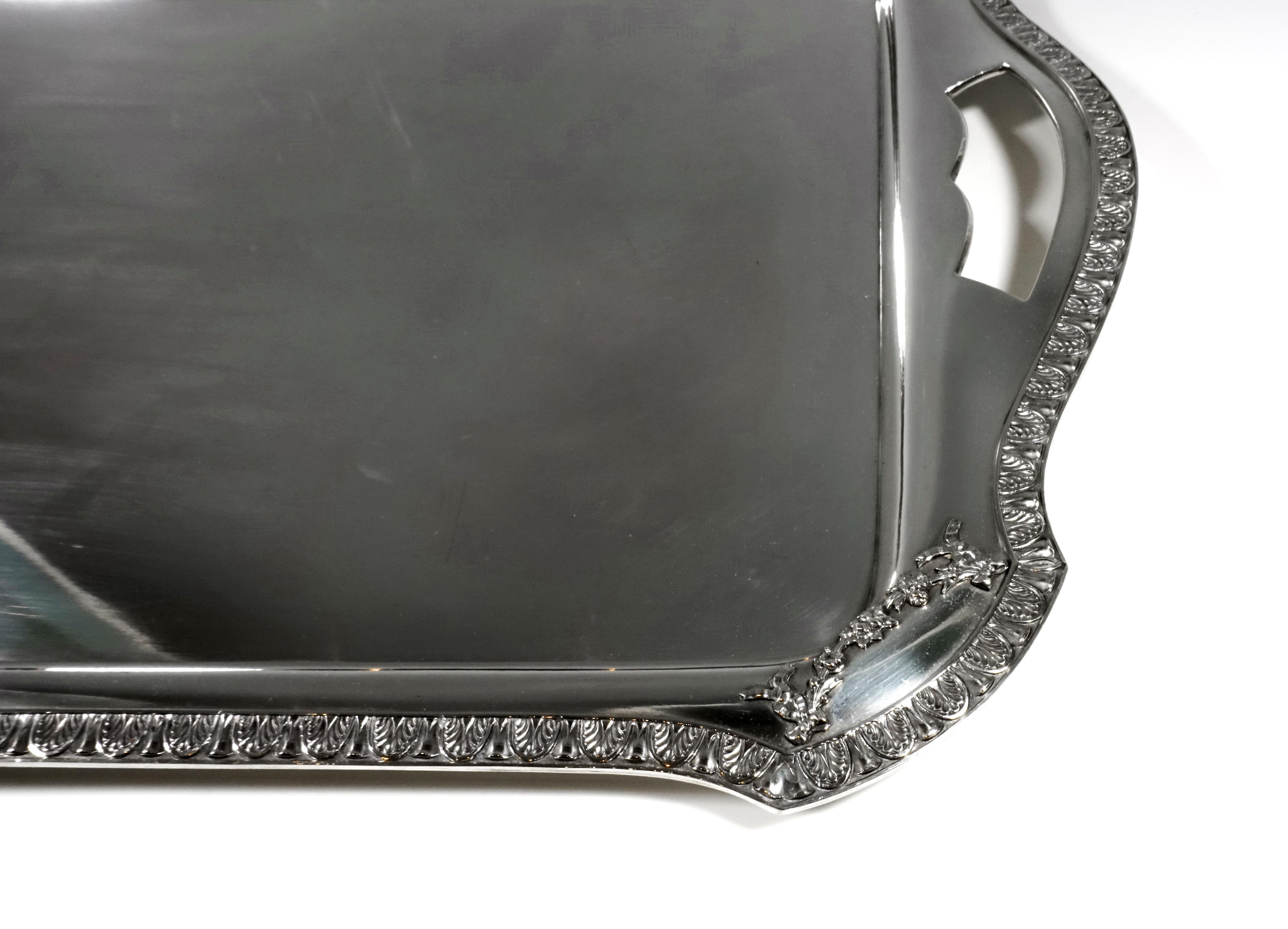 Rectangular shaped silver tray in a smooth, simple design with raised, flared edge with circumferential palmette band, beveled, rounded corners with flower garlands, extension of the edge piece on the broad sides with incised, curved
