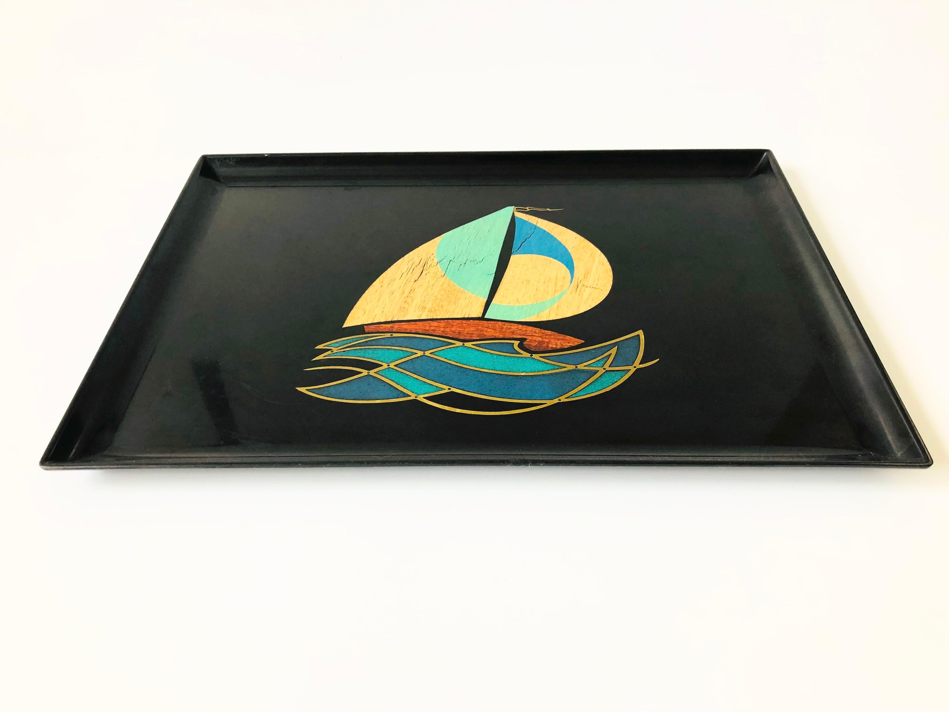 A large vintage rectangular resin tray. Small pieces of metal and wood have been inlaid into the surface to create a sailboat design. Made by Couroc.
 