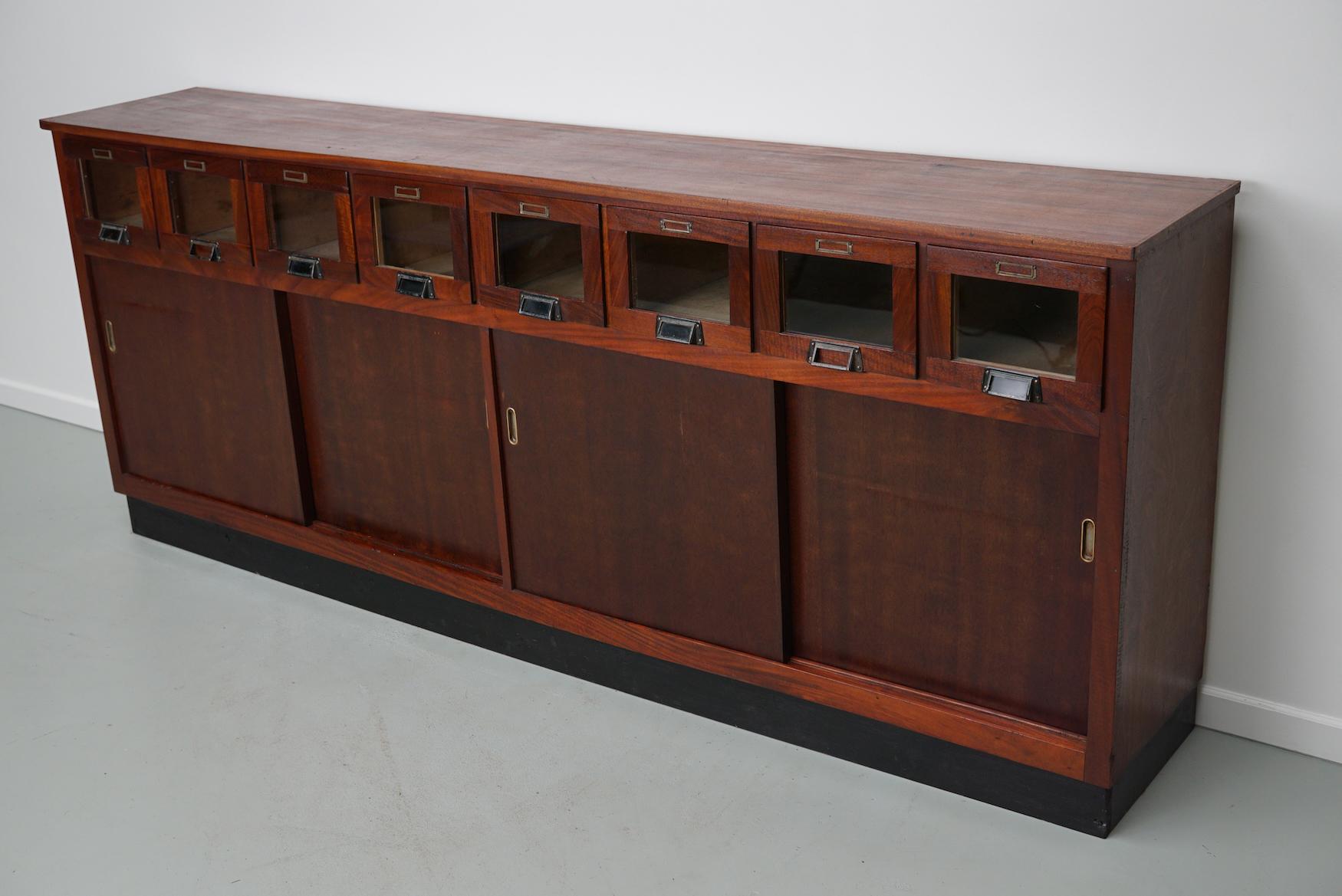 This haberdashery cabinet was produced during the 1950s in the Netherlands. It features 8 drawers in mahogany with glass fronts and black metal handles / name card holders and four sliding doors with a shelve. It was originally used in a shop for