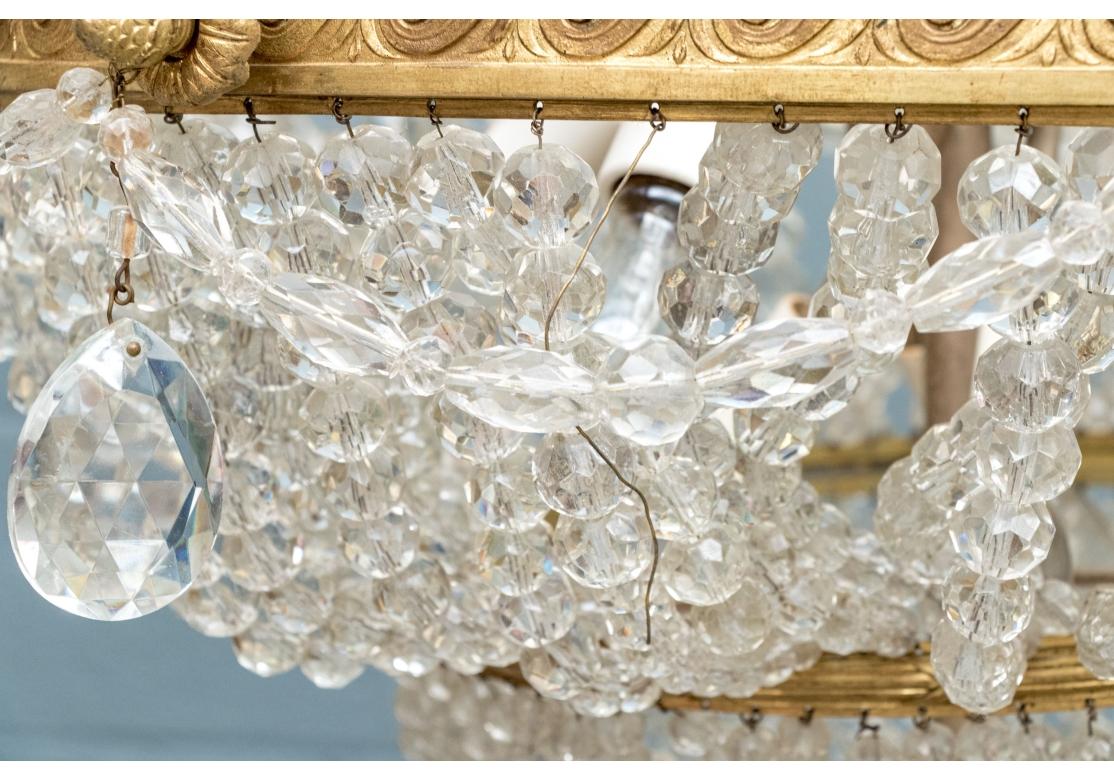 Un grande e scenografico apparecchio circolare realizzato in strati di metallo dorato con perle di vetro sfaccettate e graduate. La cornice superiore presenta una fascia dorata a forma di foglia sopra una fascia a volute e finali a carciofo