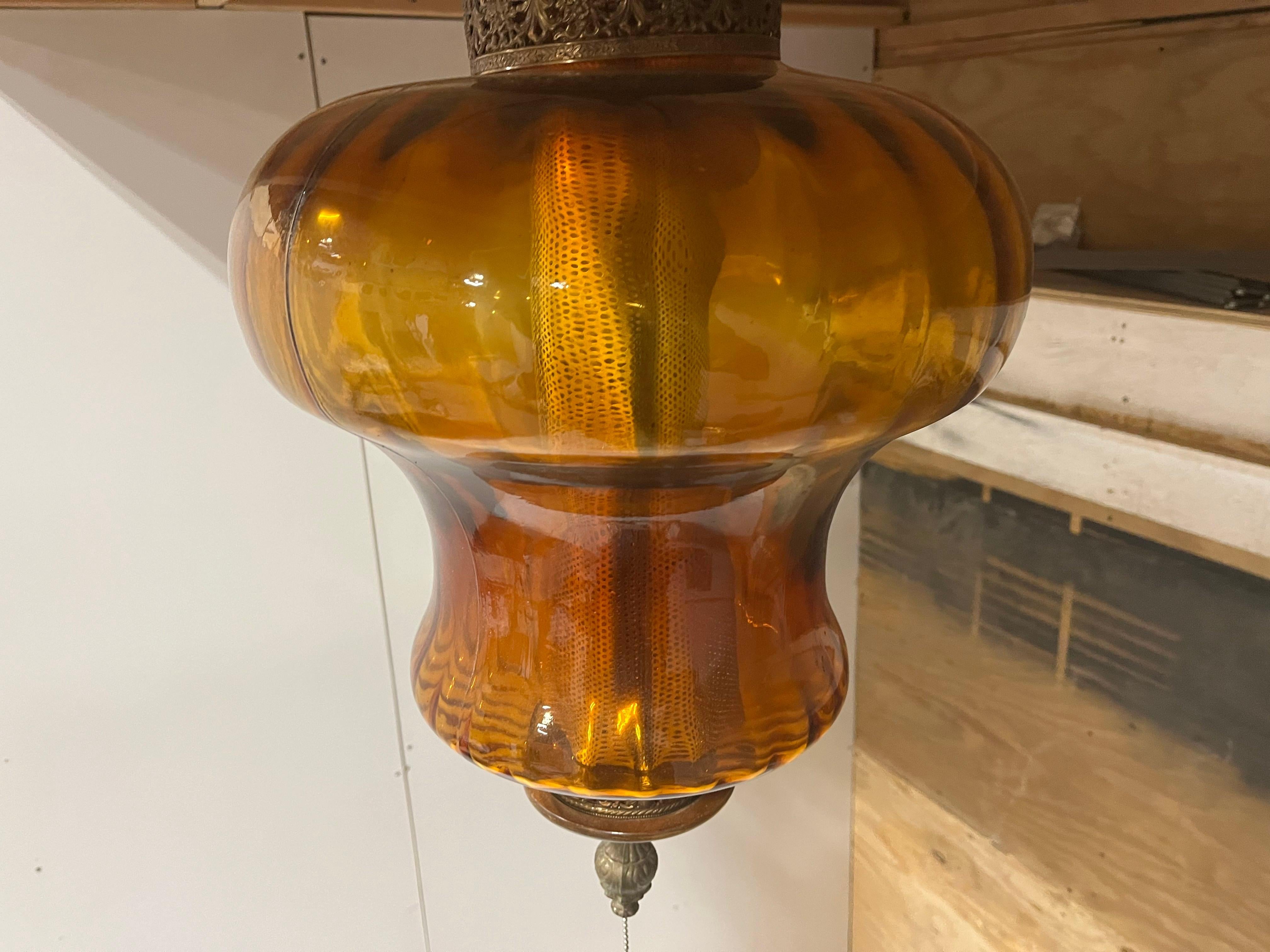 Im Mittelpunkt steht ein großzügiger bernsteinfarbener Glasschirm, der ein warmes, einladendes Licht ausstrahlt, das die Atmosphäre jedes Raumes aufwertet. Das Glas weist oft eine verzierte Textur auf, die in Verbindung mit seiner Form den optischen