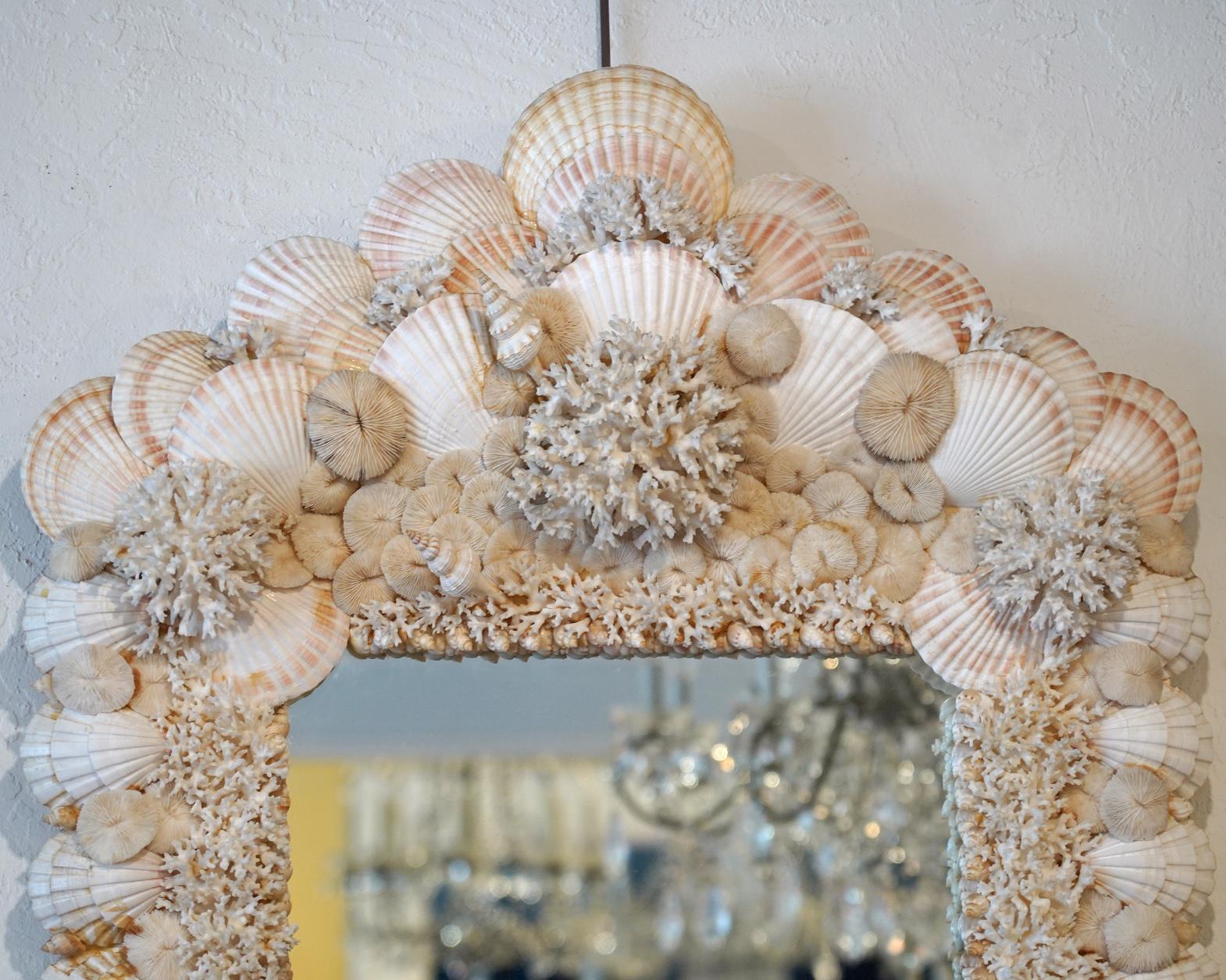 This impressing mirror is housed in a wide frame encrusted by small and large white clam shells, white branch coral and white mushroom coral composed in the manner of Christa Wilm. This mirror is a delightful sight adding a coastal flair to any