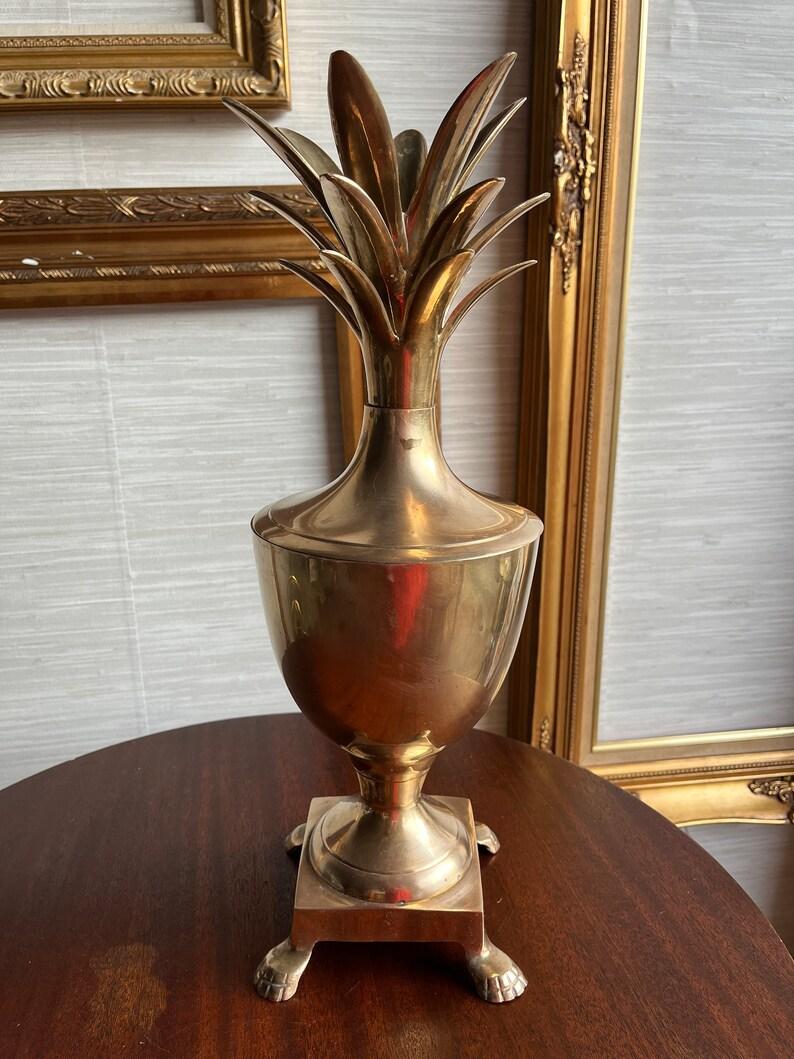 nfuse your home with timeless charm—this Mid-Century brass urn is crowned with a pineapple, long celebrated as a symbol of hospitality and warm welcome.

Expertly crafted, the lid forms a full pineapple with elegant fronds, which cleverly double as