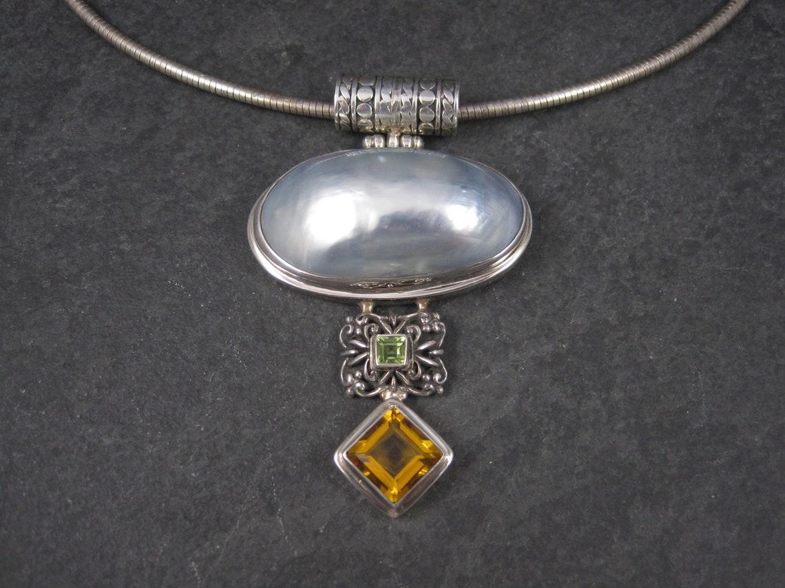 This massive 90s pendant is sterling silver.

It features a 14x42mm mabe pearl, a 5x5mm square peridot and an 11x11 square orange topaz.

This pendant measures 1 3/4 inches at its widest point and 2 3/4 inches long, including the bail.

Marks: