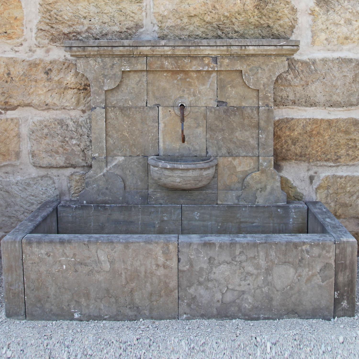 Large wall fountain with a rectangular basin (H: 48 cm) and a smaller, half-moon shaped sink. The tall rear wall shows a slightly protruding cornice and a filling with rounded corners. The fountain is hand-carved out of bluestone.