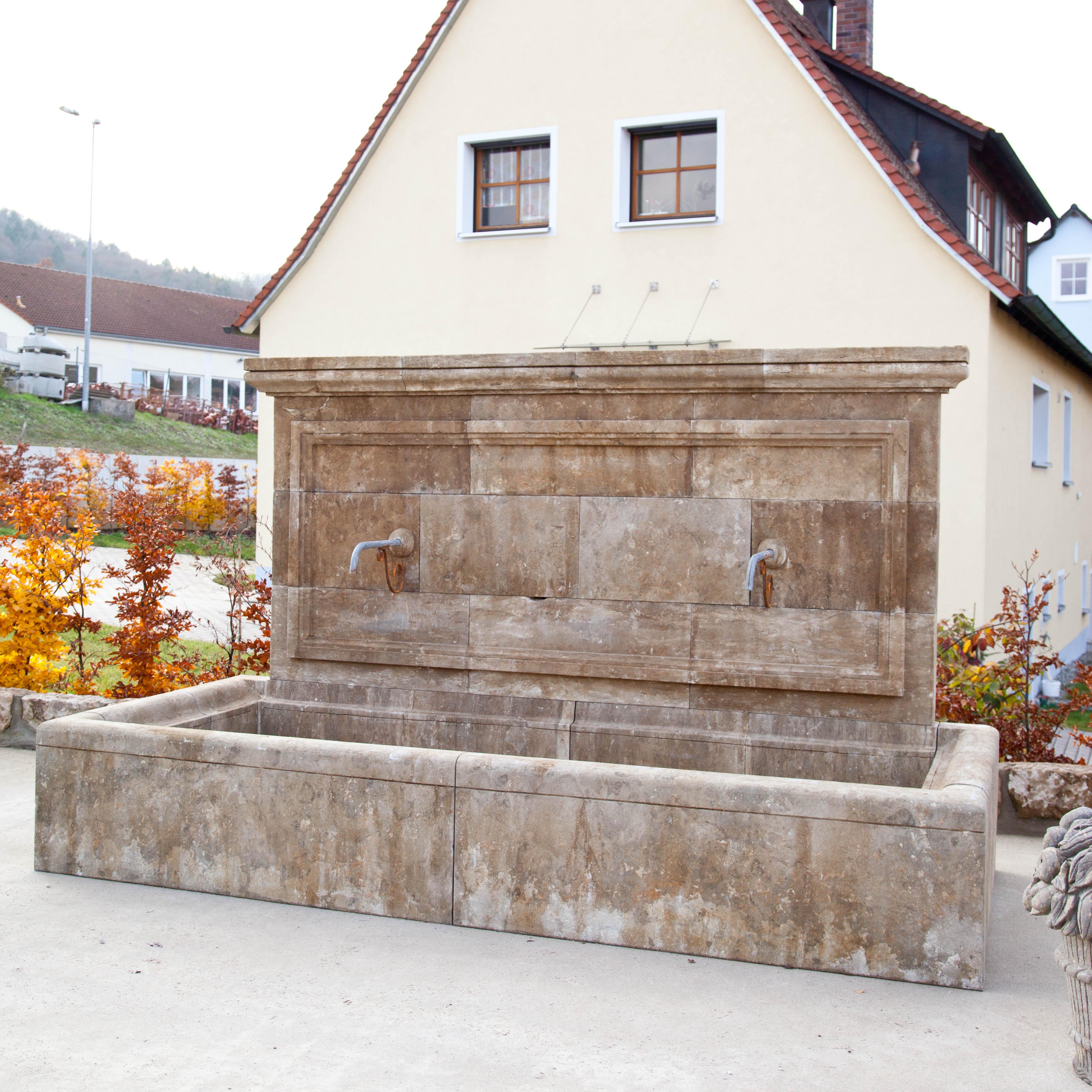 Large blue stone wall fountain with rectangular basin. The back wall is architecturally structured and equipped with two spouts. Measures: Basin height 50 cm.