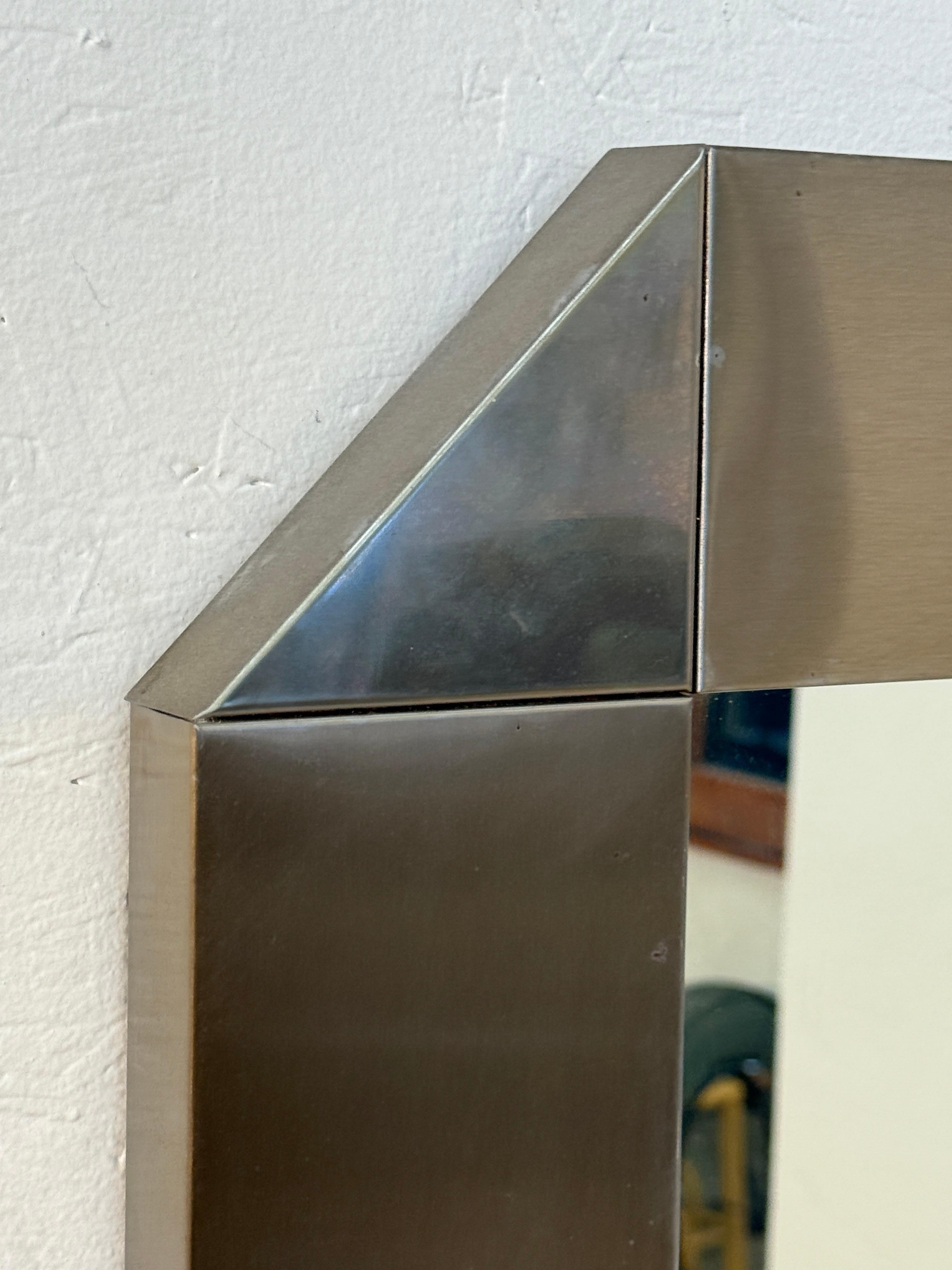 Ce miroir mural contemporain est doté d'un cadre large et audacieux en métal brossé. Le design épuré et géométrique est accentué par les bords biseautés en angle, créant profondeur et dimension tout en reflétant la lumière avec une élégance moderne.
