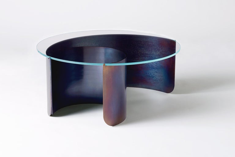 Rolled sheets of steel curl and peel away beneath a transparent glass surface, capturing the dynamic moment of a cresting wave. The vibrant multicolored steel surface is created through a unique heat treatment process through which molecules realign