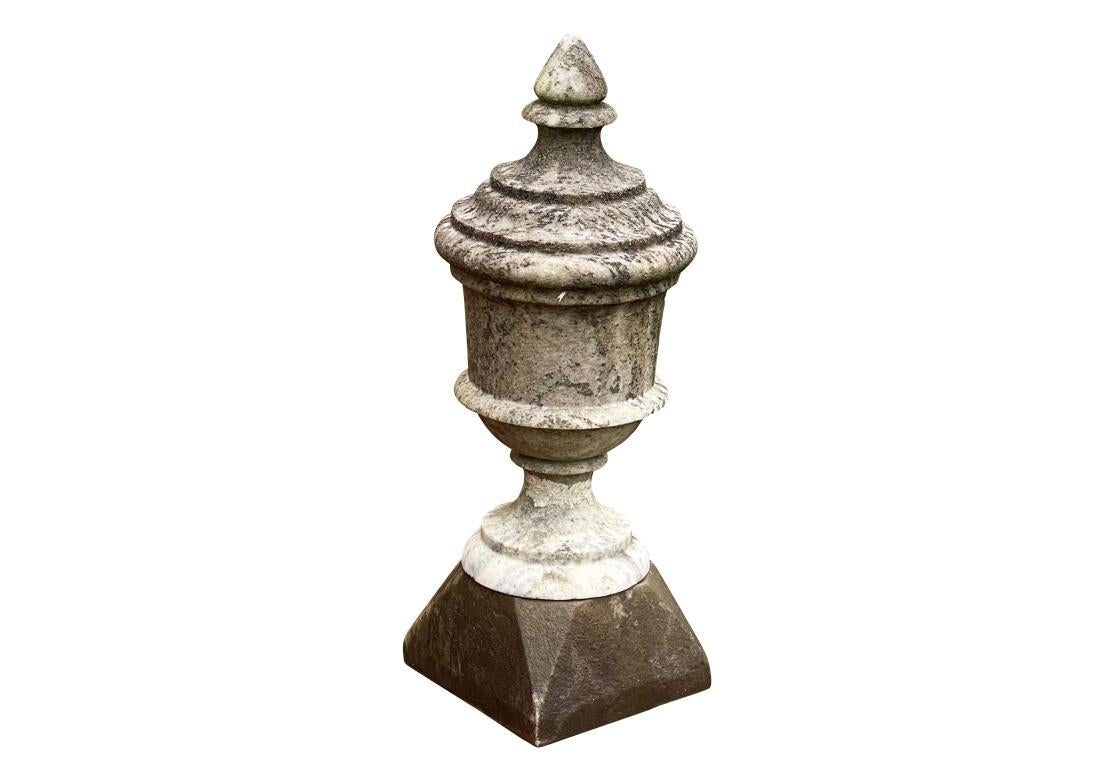 Grande urne en marbre de forme traditionnelle. La partie supérieure de l'urne a pris une teinte grise plus foncée, tandis que la partie inférieure, protégée, a conservé sa teinte claire d'origine. L'urne est montée sur un socle dont la teinte est