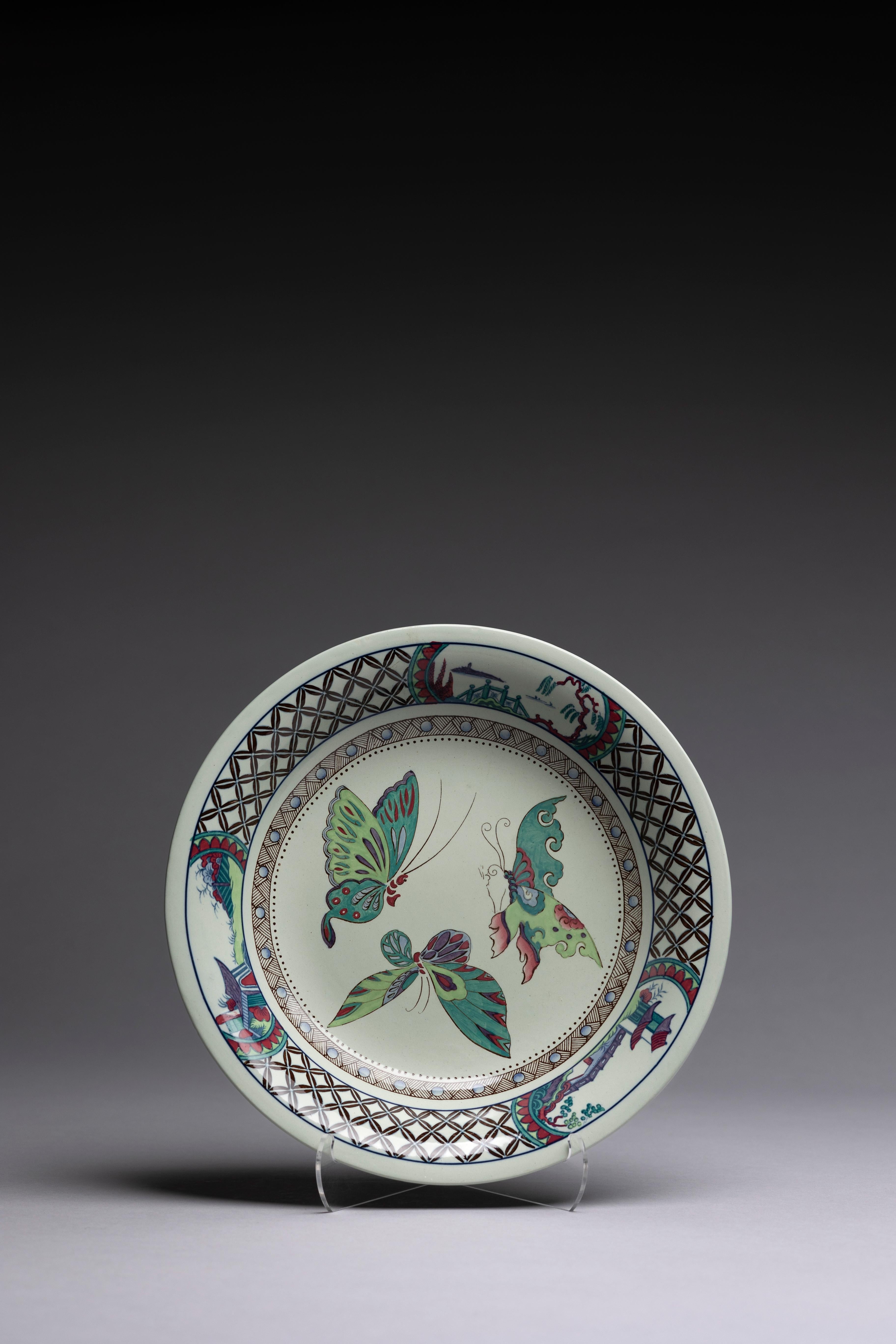 Eine flache Servierschale mit bunten Schmetterlingen, hergestellt von Wedgwood um 1915.

Drei lebhafte Schmetterlinge tanzen um die Mitte dieser Schale, die von einem Rand im orientalischen Stil mit Vignetten von fantastischen asiatischen
