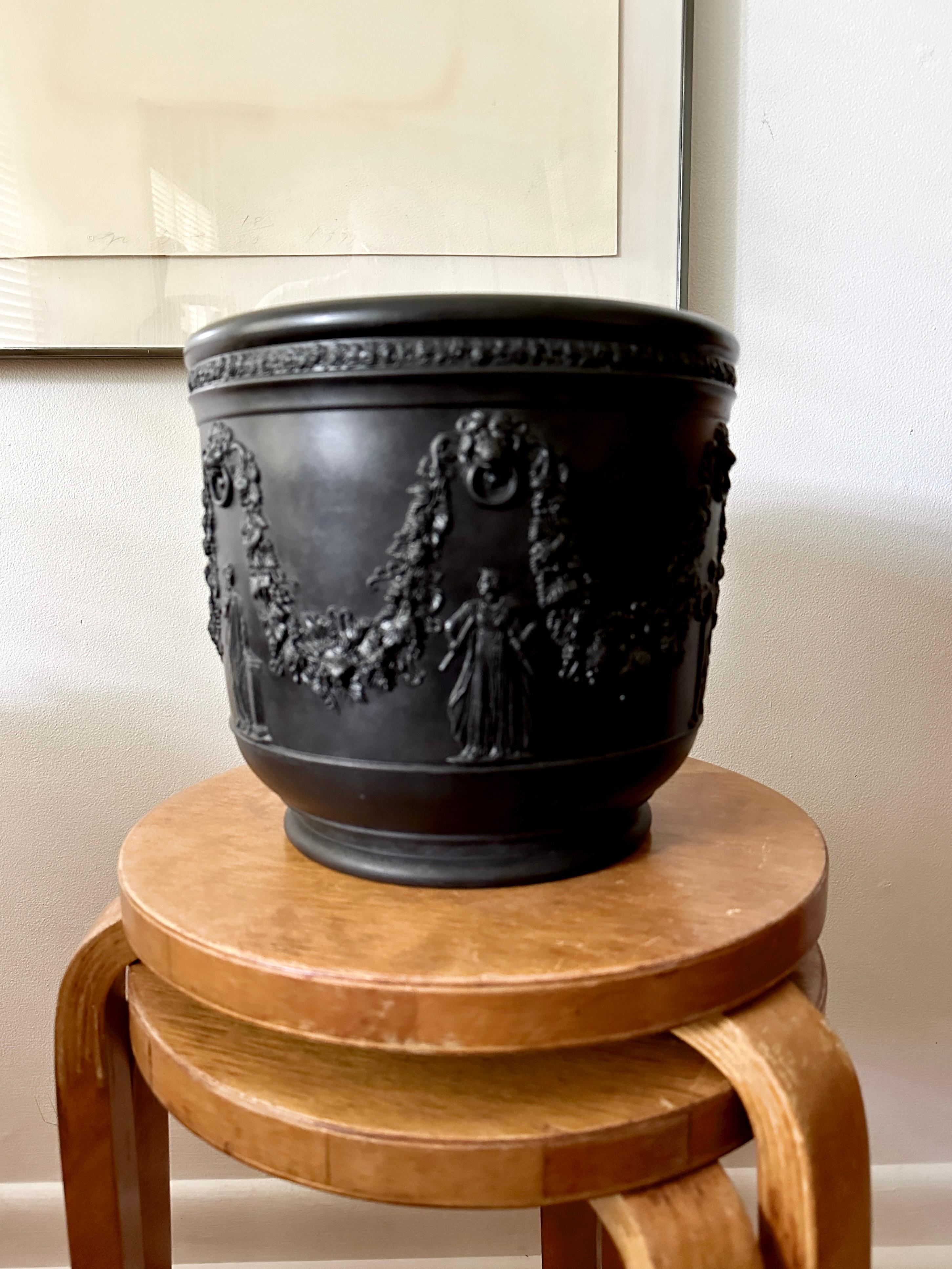Giardiniera in basalto nero di Wedgwood, realizzata in Inghilterra intorno al 1900. La giardinetta ha un bordo a fasce floreali sopra festoni di vite fruttiferi che terminano con maschere di leone e anelli con le Muse in basso. Raro formato grande