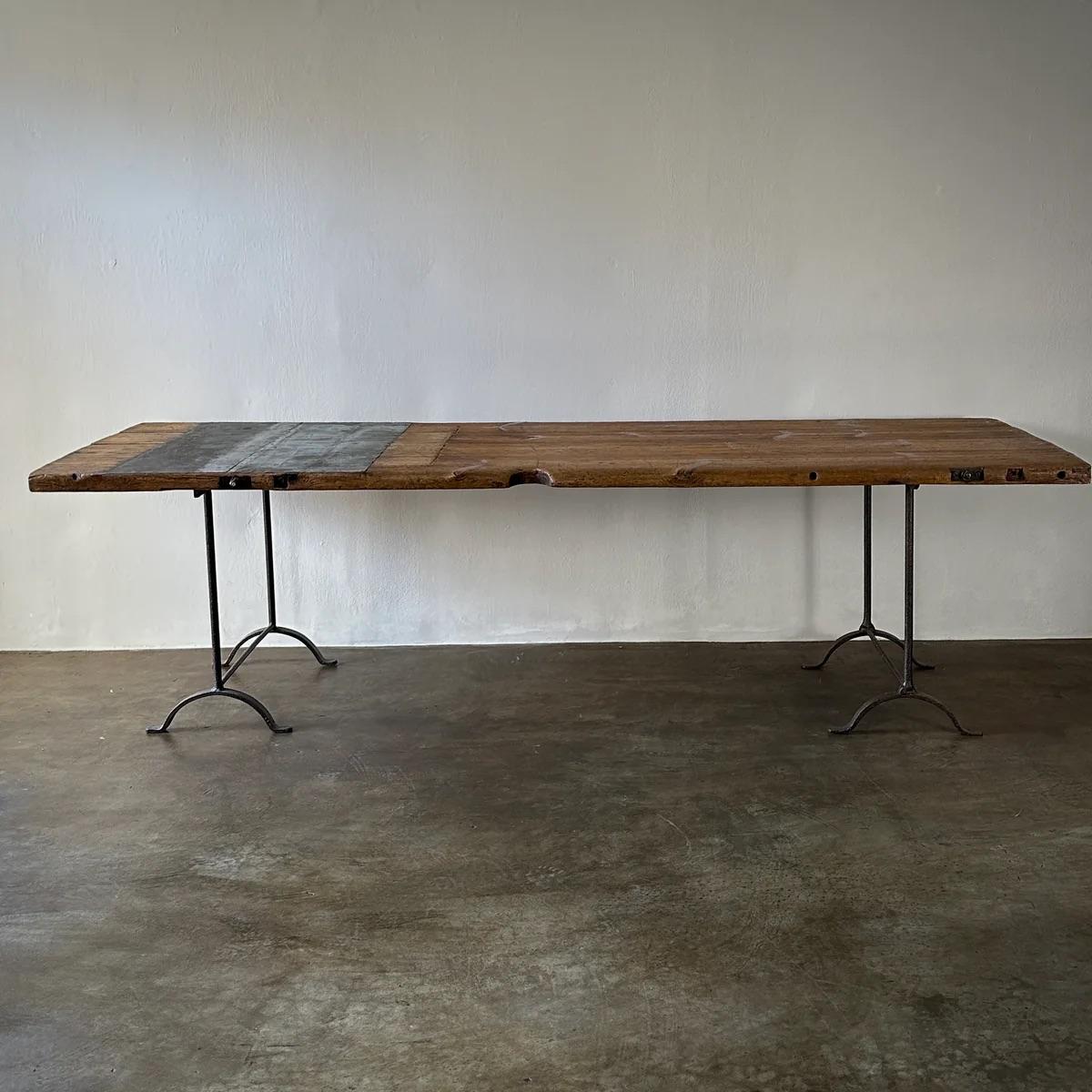This expansive wood jeweler's work table combines robust functionality with visual elegance. Mounted on a sturdy iron trestle base, the dense pine top showcases a deep, inviting patina, reflective of its rich history in craftsmanship. Its ample size