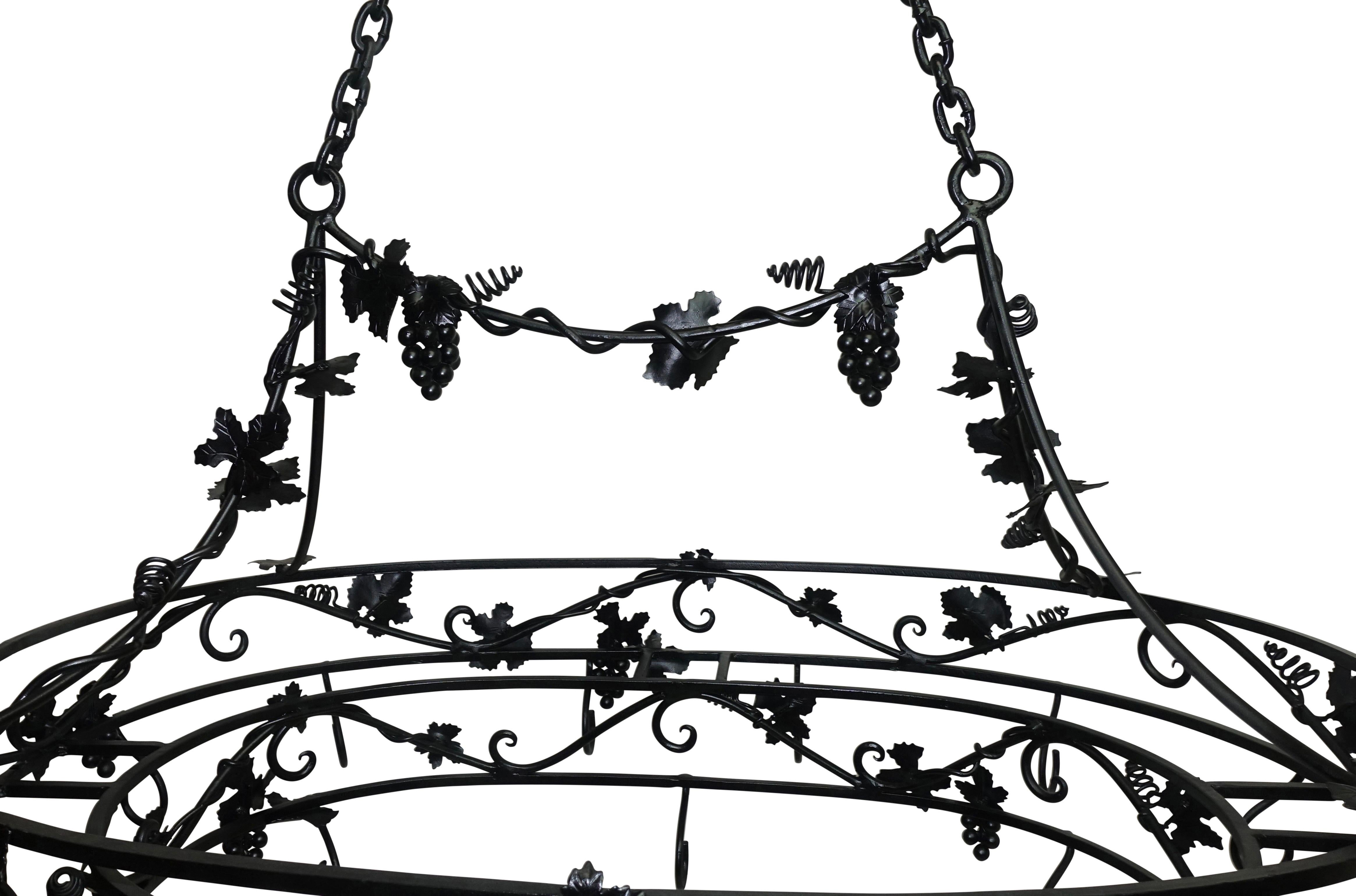 A large oval shape wrought iron hanging pot rack with clusters of grapes and leaves. Having two tiers of hanging hooks, the lowest tier with six stationary hooks and the larger oval tier with twelve stationary hooks, suspended from a double brass